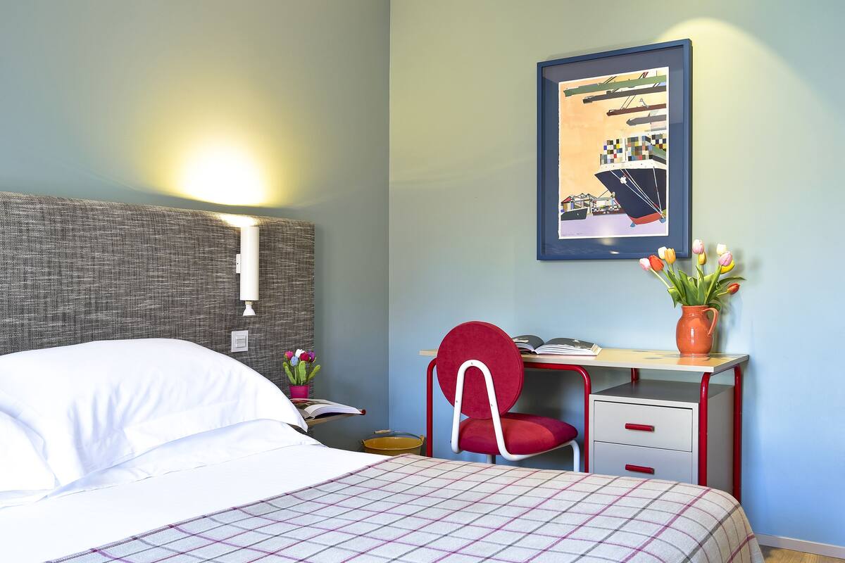 A neatly arranged hotel room with a bed, a desk with a chair, a framed picture on the wall, and a vase with flowers on the desk.