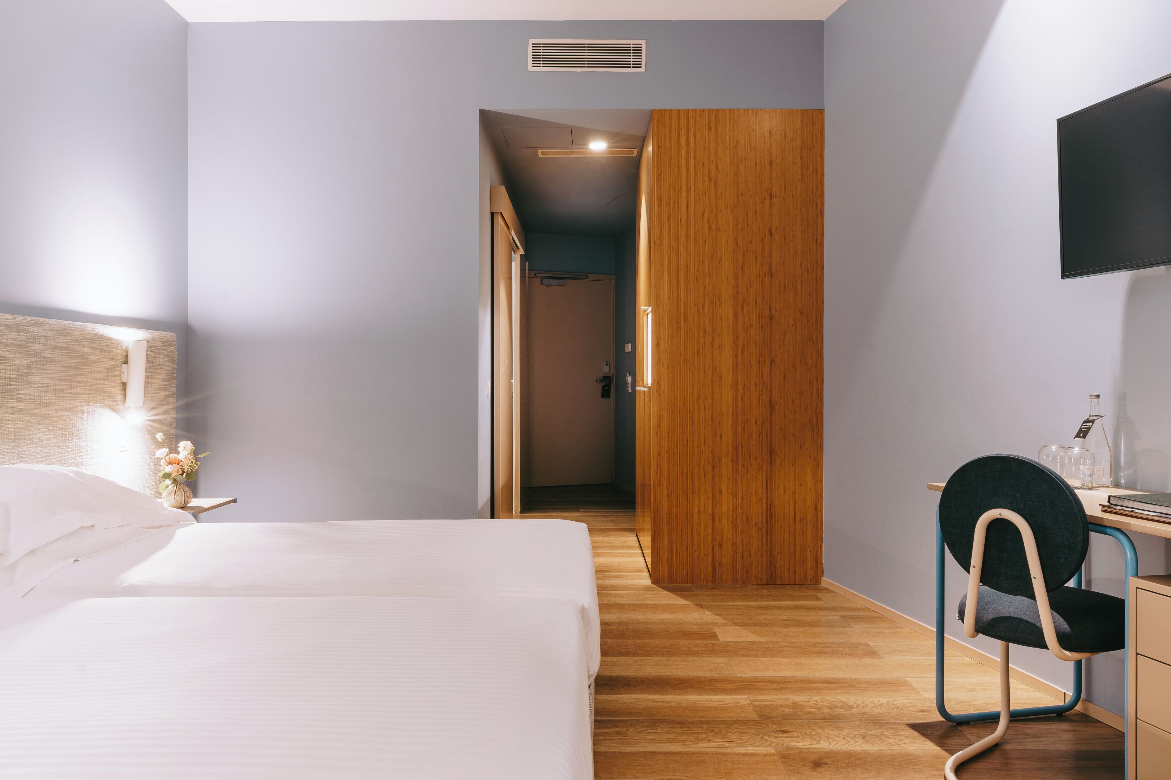 A hotel room with a bed, a desk with a chair, a television, and a wooden wardrobe.