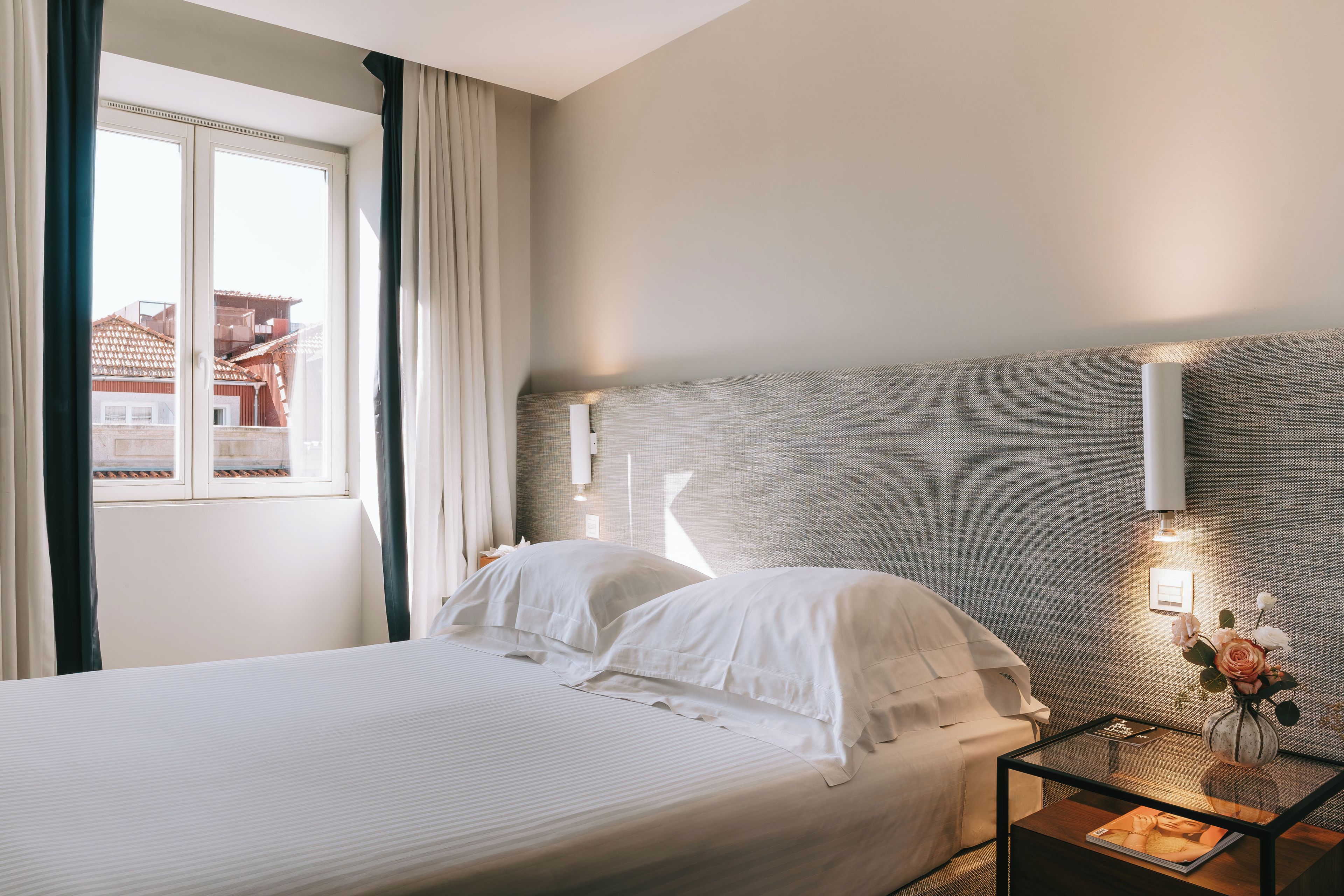 A hotel room with a bed, a nightstand, a vase with flowers, and a window with curtains.