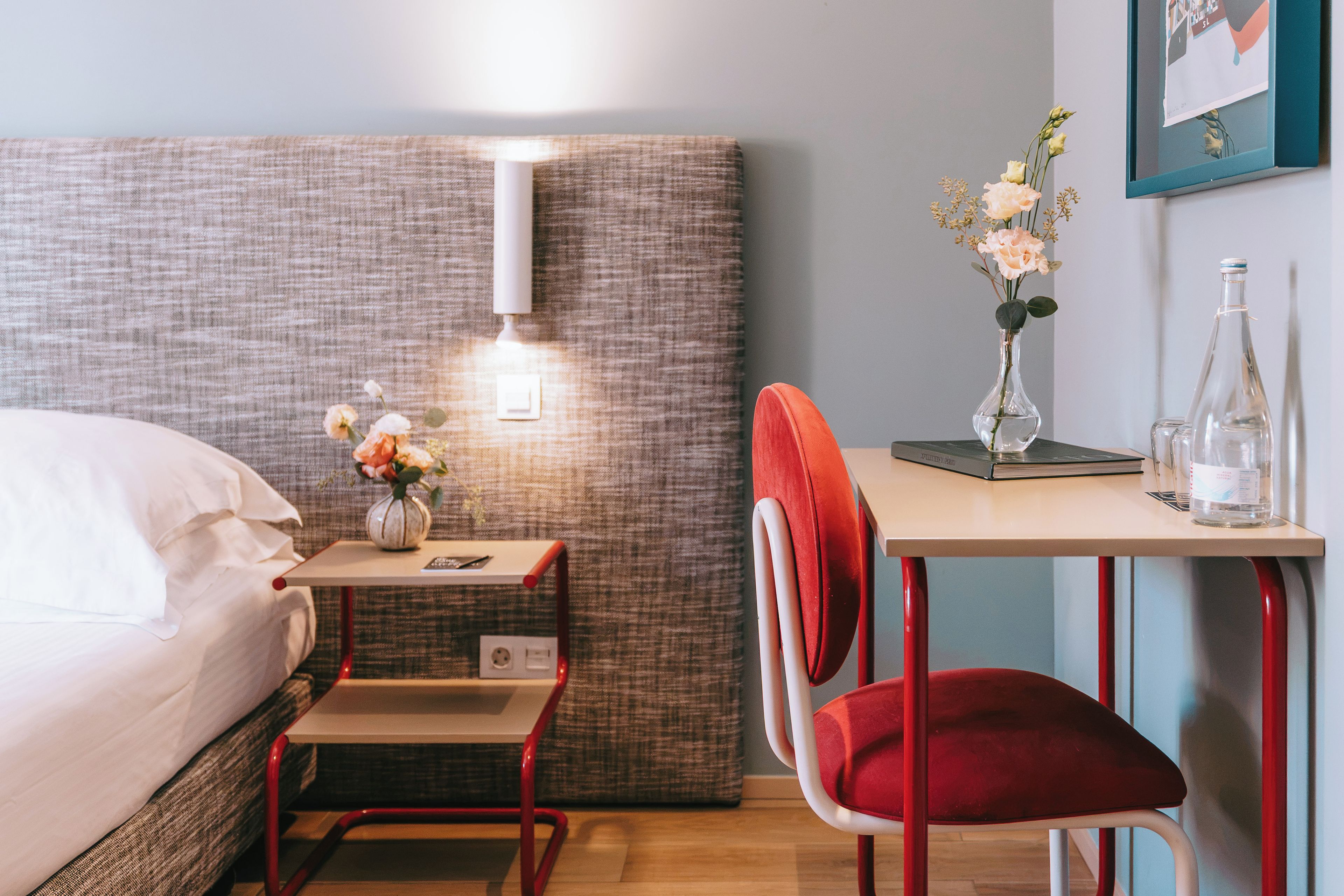 A hotel room with a bed, a desk with a chair, a vase with flowers, and a bottle.