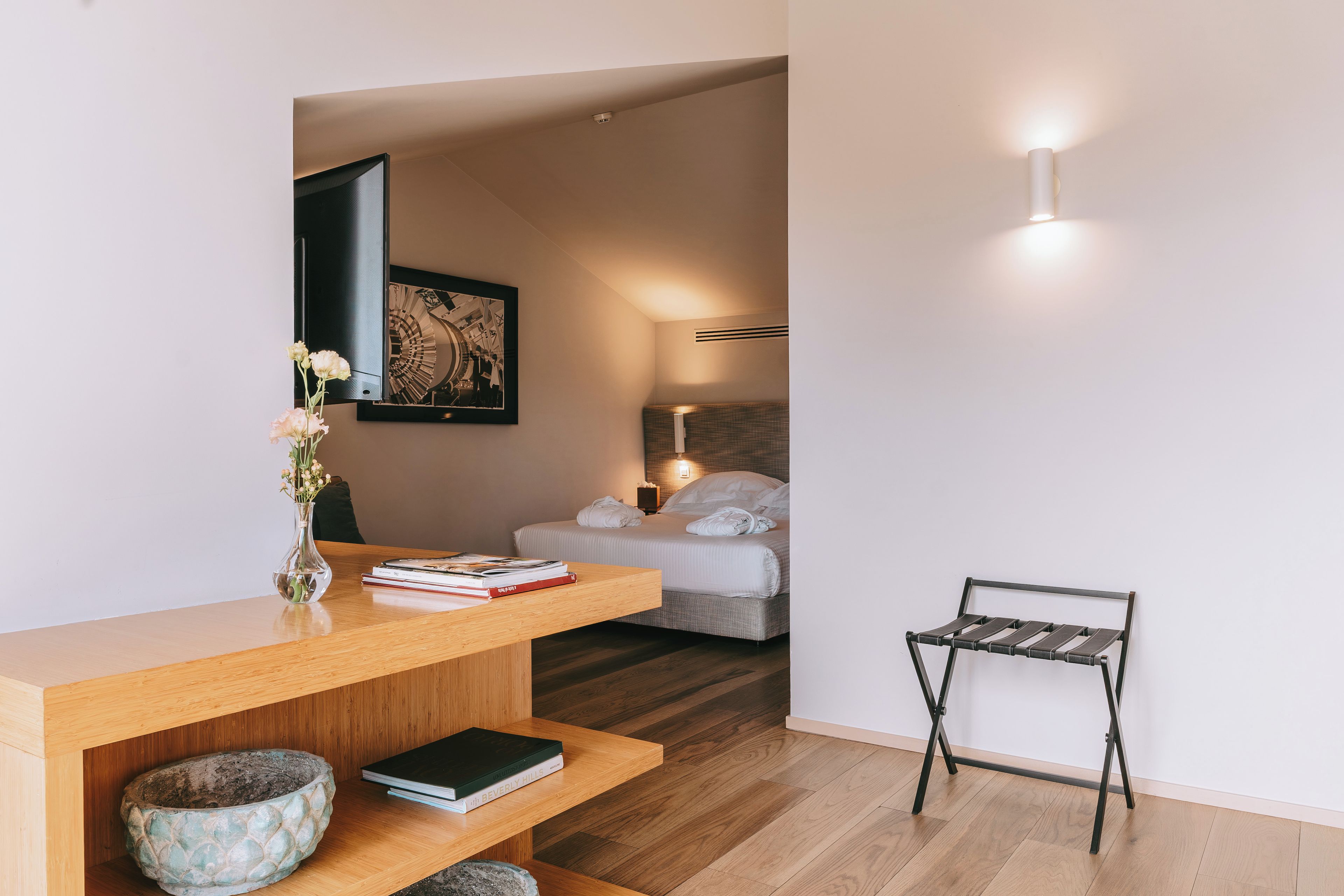 A modern bedroom with a wooden desk, a bed with white bedding, a small table, and a wall-mounted light fixture.