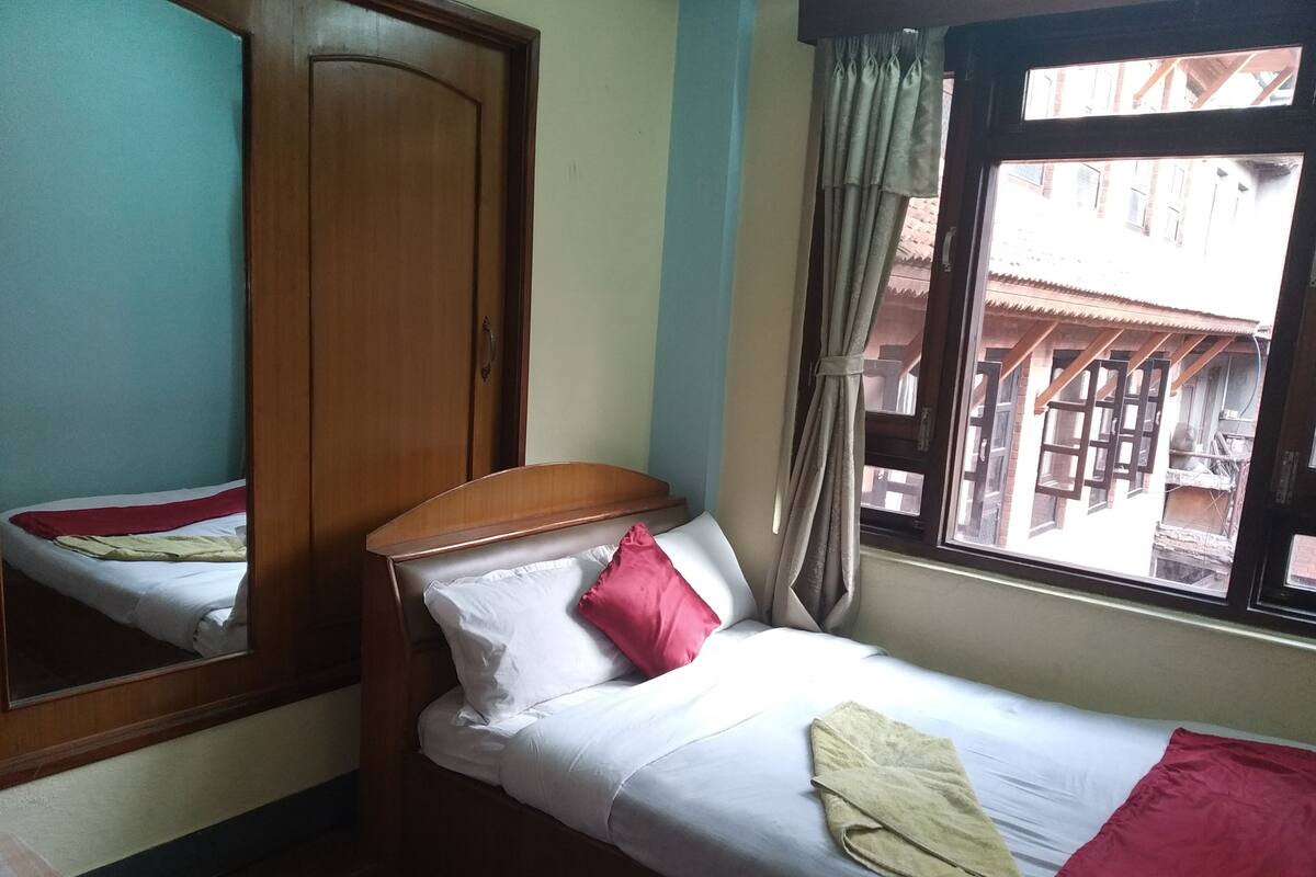 Ein Einzelbett mit weißer und roter Bettwäsche, ein Holz-Kopfteil, ein Spiegel, ein Fenster mit Vorhängen und Blick auf Gebäude.