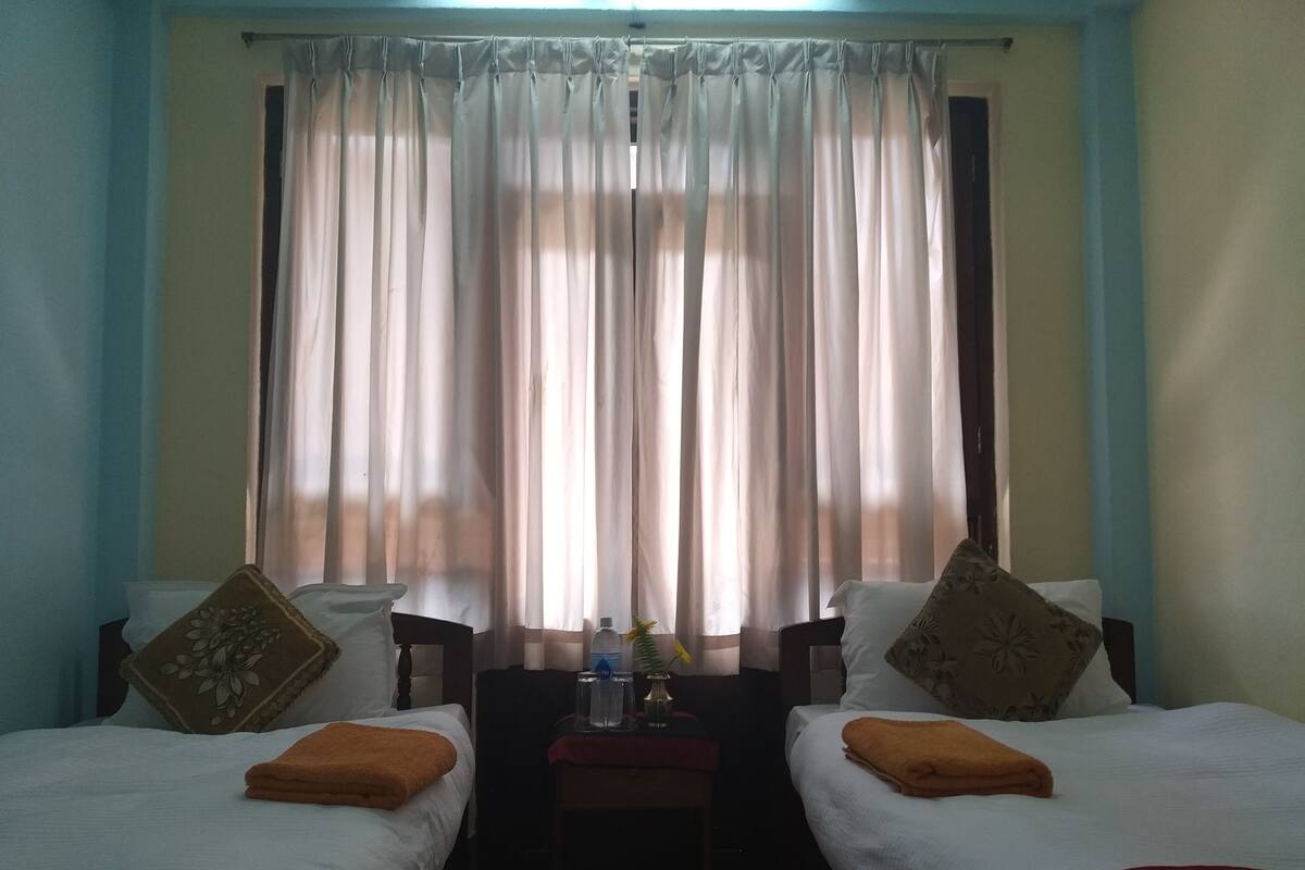 Ein Hotelzimmer mit zwei Betten, einem kleinen Tisch mit einer Flasche und einer Pflanze sowie einem Fenster mit transparenten Vorhängen.