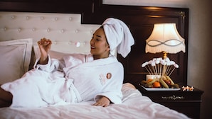 A person in a white bathrobe with a towel on their head, lying on a bed with a tufted headboard. A bedside table with a lamp and a fruit platter is visible. - Latanya Palm & Spa Airport Antalya (Antalya)