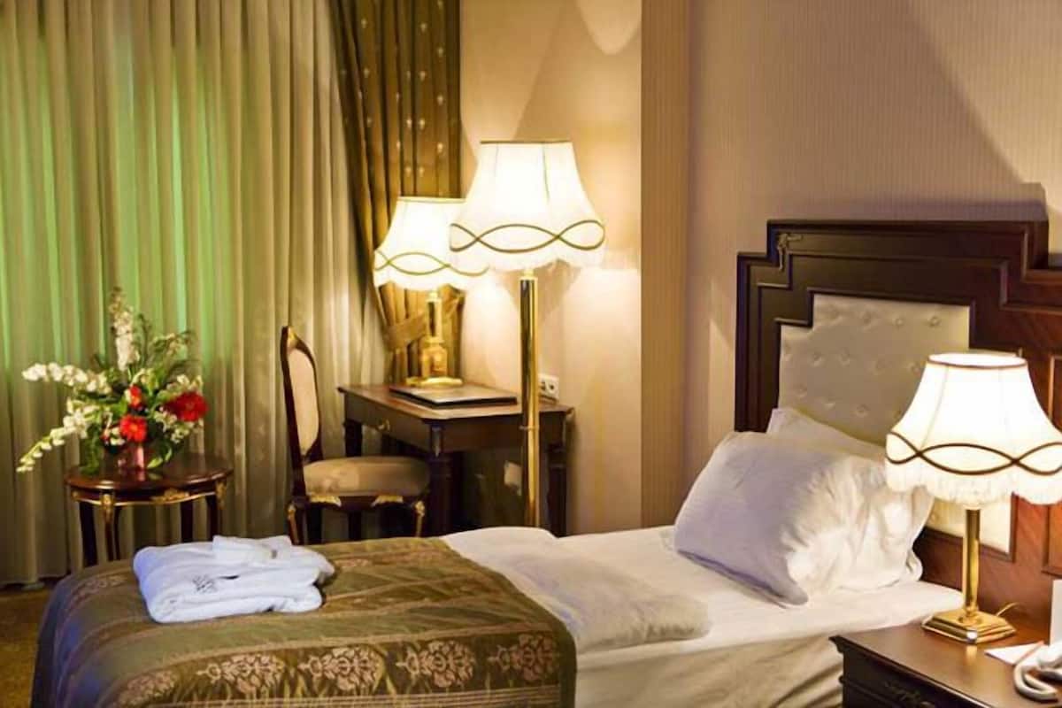 Ein Hotelzimmer mit einem ordentlich bezogenen Bett, zwei Nachttischlampen, einem kleinen Tisch mit Lampe, einem Sessel und einer Vase mit Blumen.