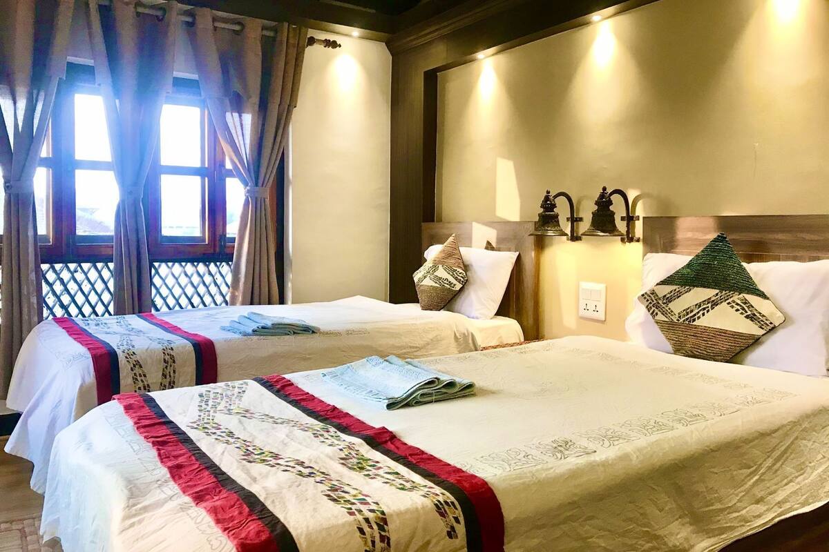 A hotel room with two beds, a wooden headboard, wall-mounted lights, and a window with curtains.