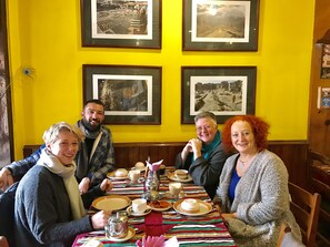 Un grupo de personas sentadas en una mesa con mantel a rayas, cuadros en la pared y una decoración con pluma de pavo real.