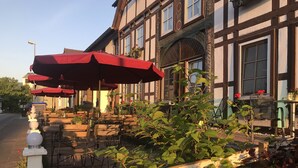 En udendørs café med træborde og røde parasoller, omgivet af grøn vegetation og en traditionel bygning med store vinduer.