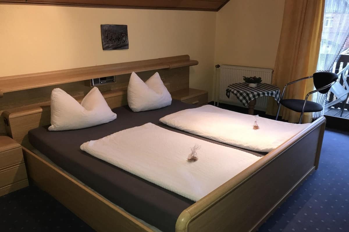 Uma cama de casal com duas almofadas, uma mesa de cabeceira, uma cadeira e uma pequena mesa com toalha xadrez.