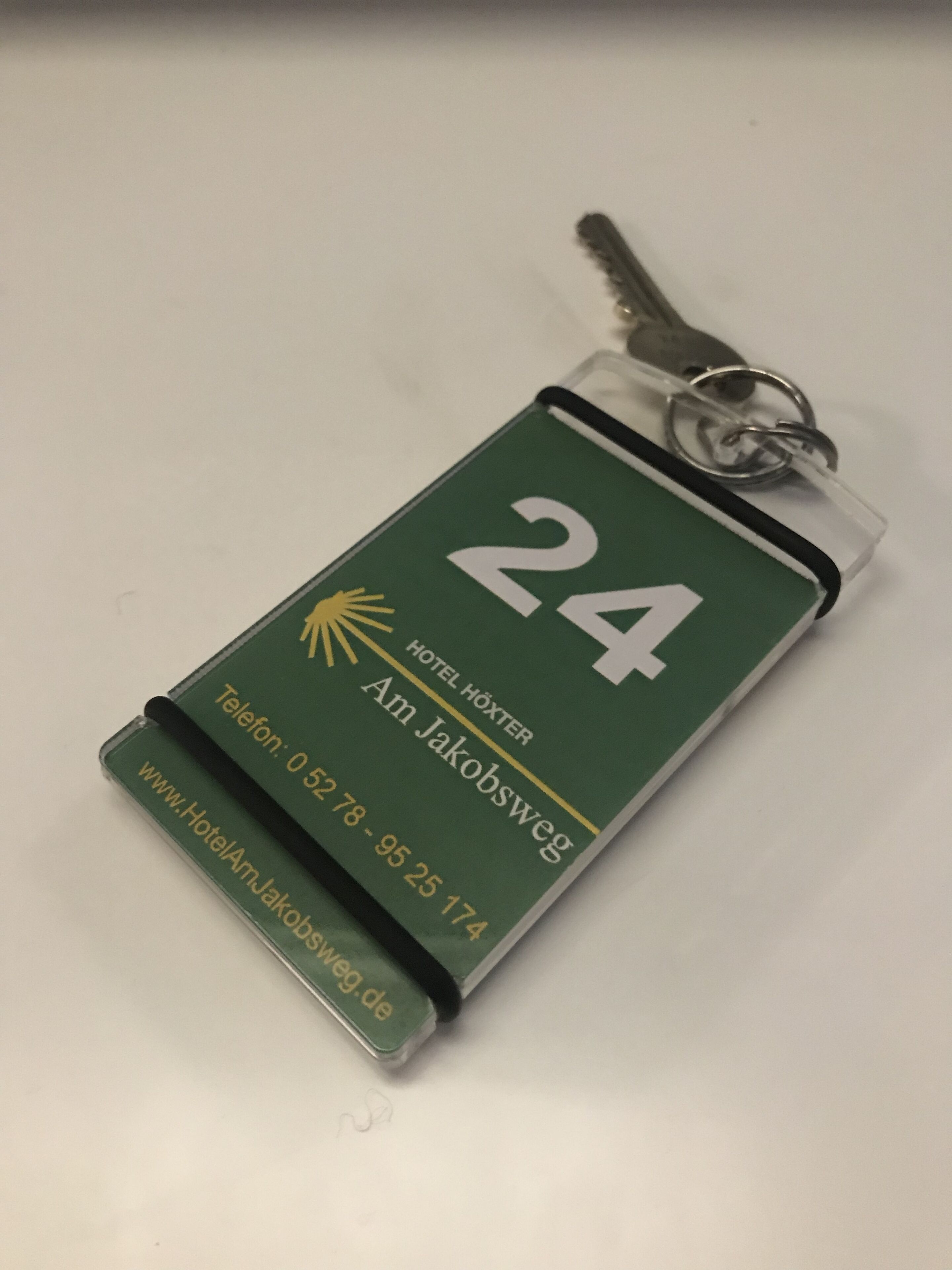 Een sleutelhanger met een groene achtergrond, het cijfer 24 en tekst met 'Hotel Høxter, Am Jakobsweg, Telefoon: 05278-95 25 174, www.HotelAmJakobsweg.de'.