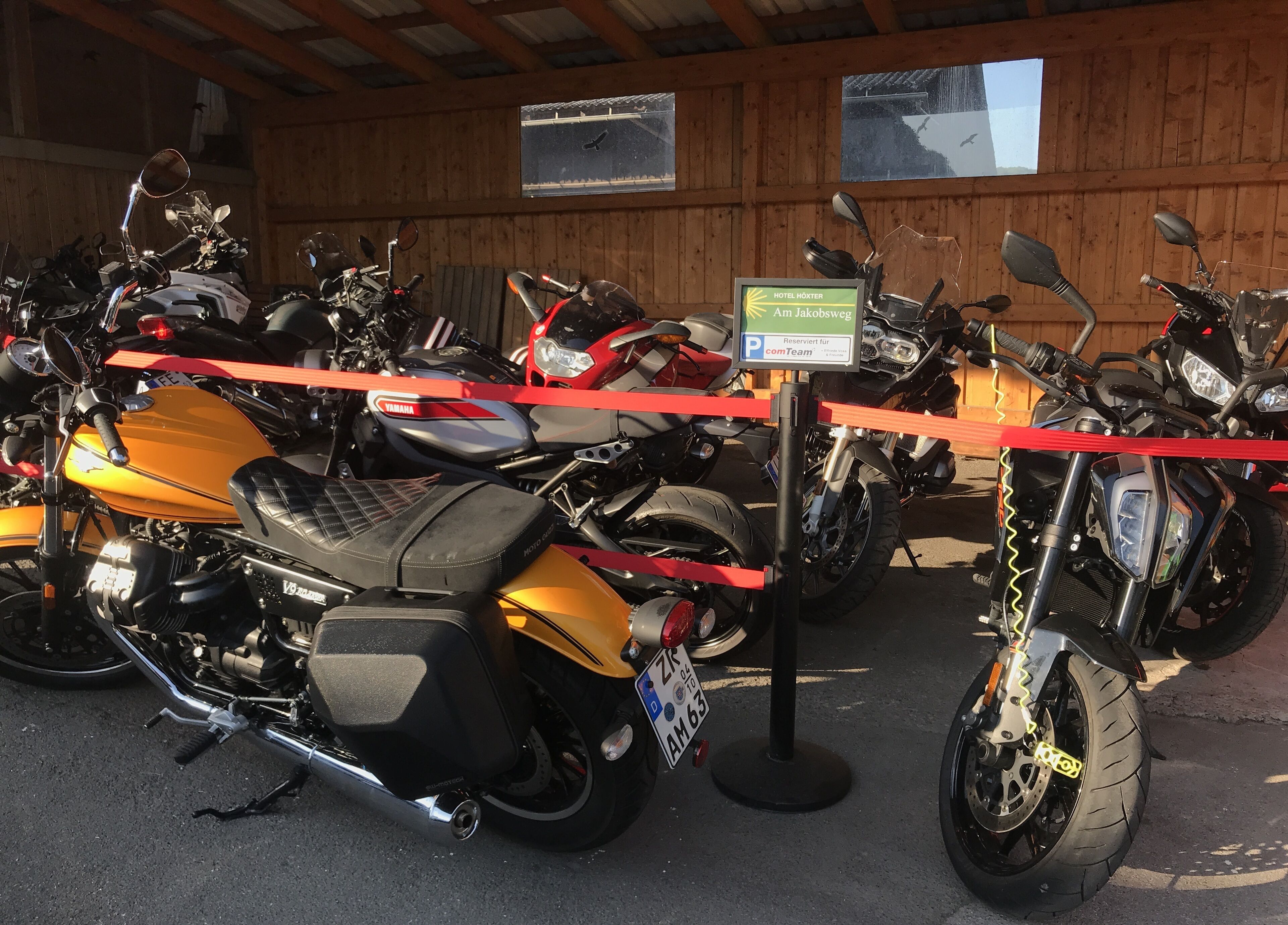 Een rij motorfietsen geparkeerd in een overdekte ruimte met houten wanden en een bord waarop een hotel wordt aangegeven.