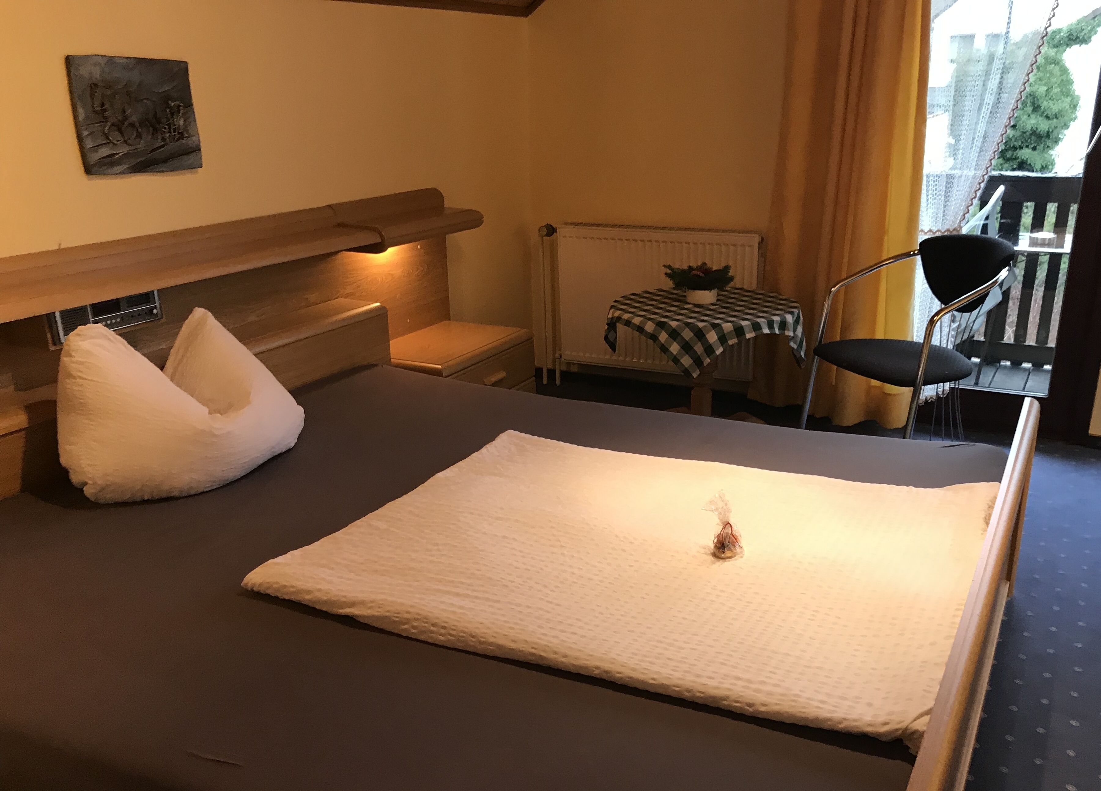 Quarto de hotel com cama, uma cadeira, mesa com toalha xadrez e janela com cortinas.