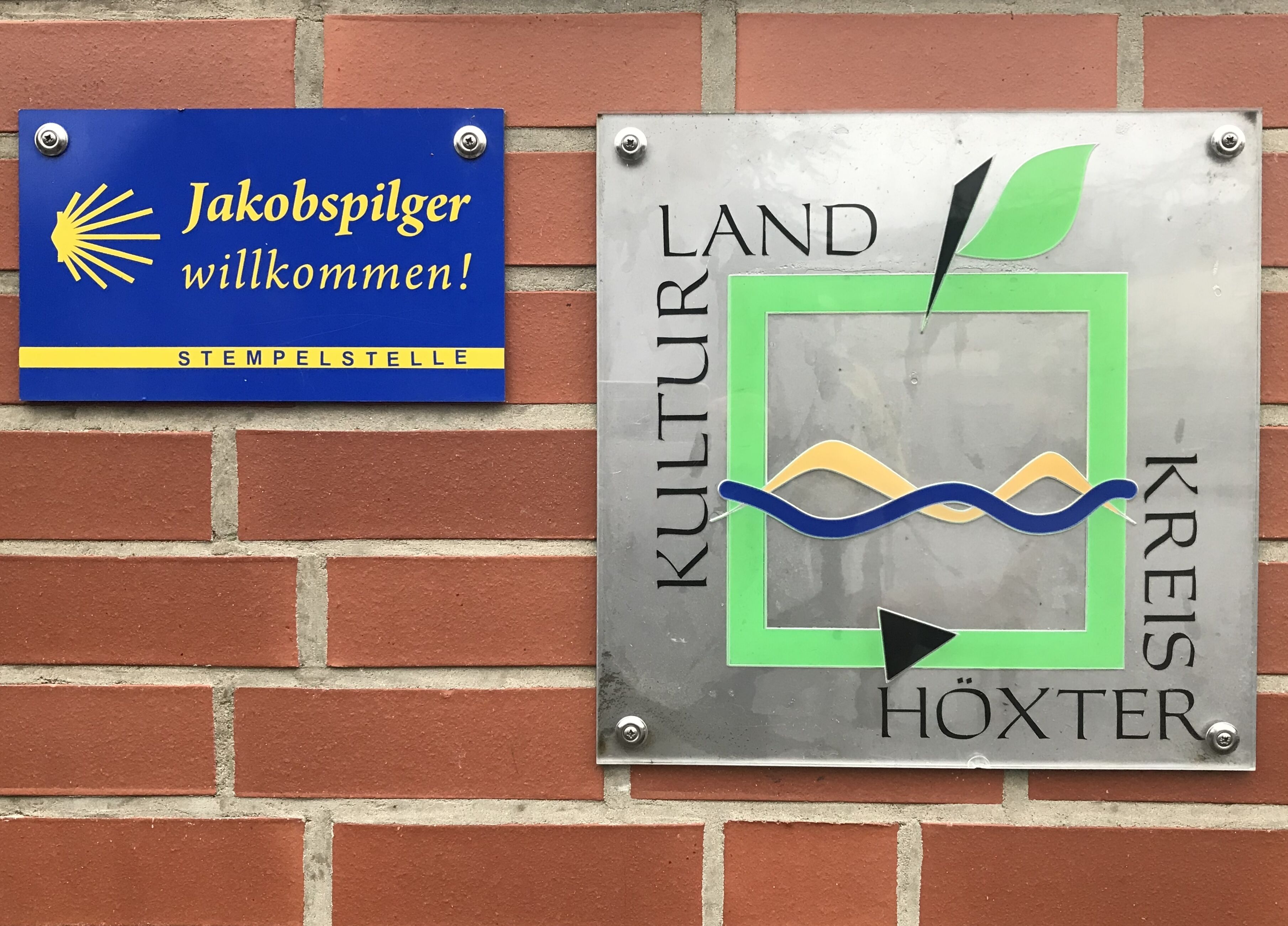 Two signs mounted on a brick wall. One sign is blue with yellow text and a sun symbol, reading "Jakobspilger willkommen! Stempelstelle." The other is silver with green and black text, reading "KULTUR AND KREIS Höxter."