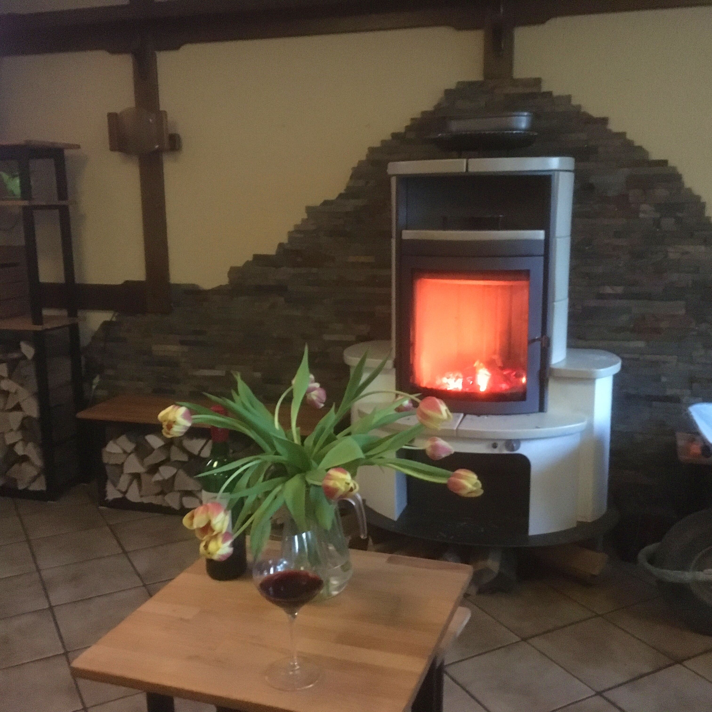 A cozy room with a stone fireplace, a table with a vase of flowers and a glass of red wine, and a stack of firewood.
