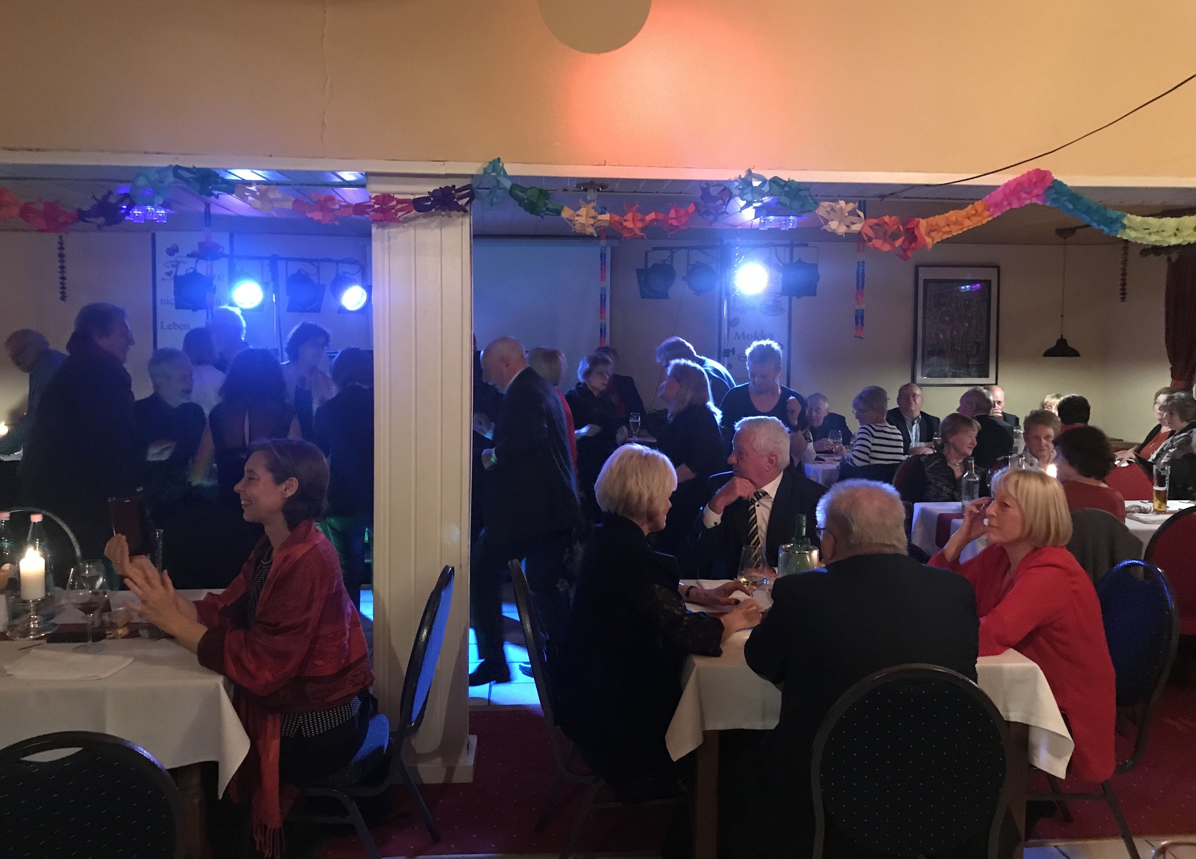 Ein lebhaftes Esszimmer mit Tischen und Stühlen, Personen sitzen und stehen, und eine festliche Dekoration mit Papierblumen und Bändern.