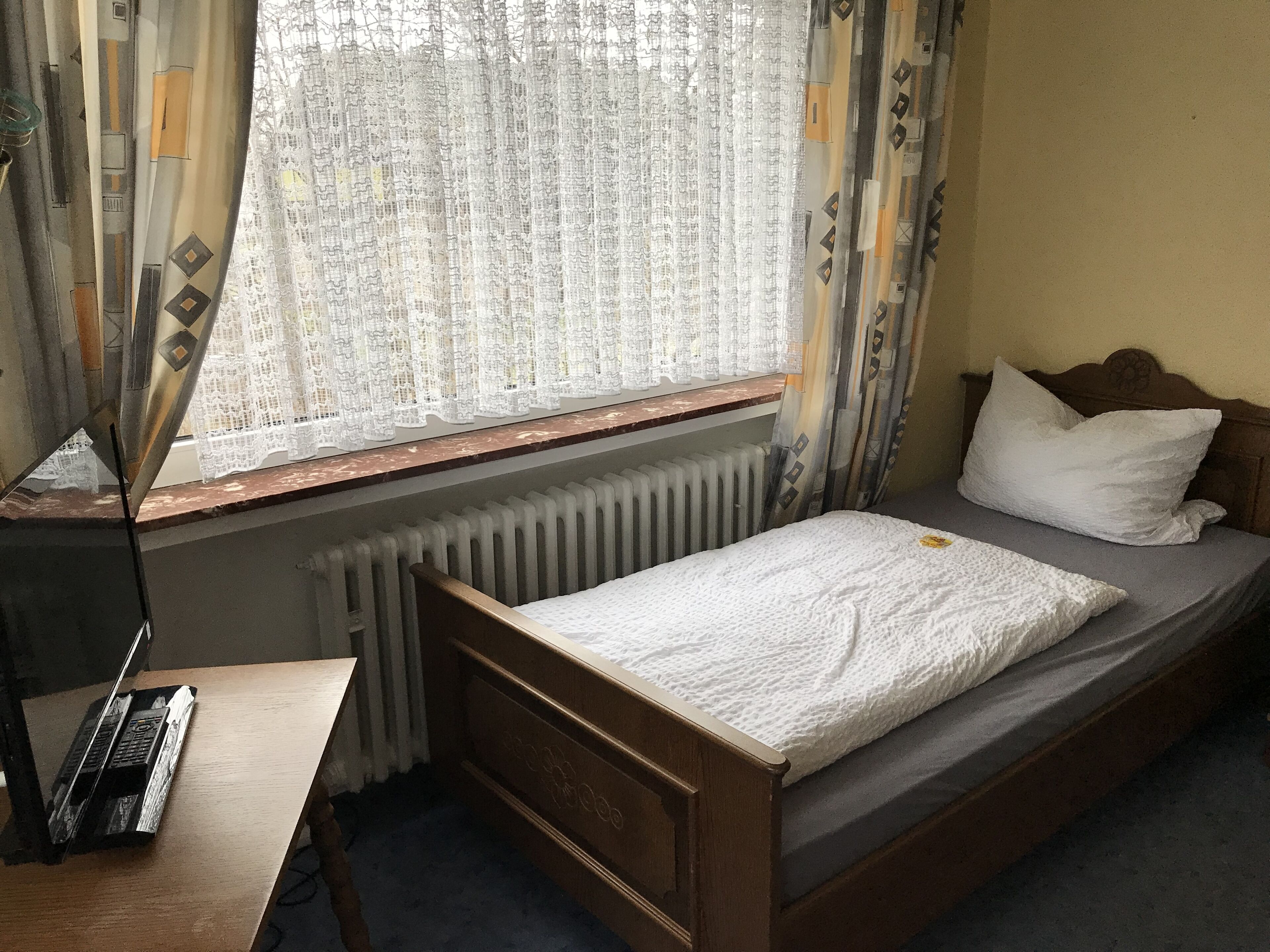 Una cama individual con cabecera de madera, una mesita de noche, un radiador y una ventana con cortinas estampadas.
