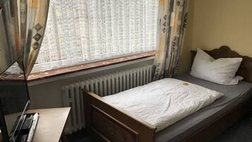 Una cama individual con cabecera de madera, una mesita de noche, un radiador y una ventana con cortinas estampadas.