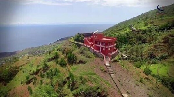 A red building with a spiral staircase leading to the entrance, surrounded by lush green hills and overlooking a body of water.