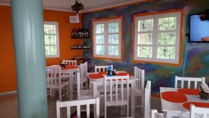 Zona pranzo con tavoli e sedie bianche, pareti arancioni e ampie finestre.
