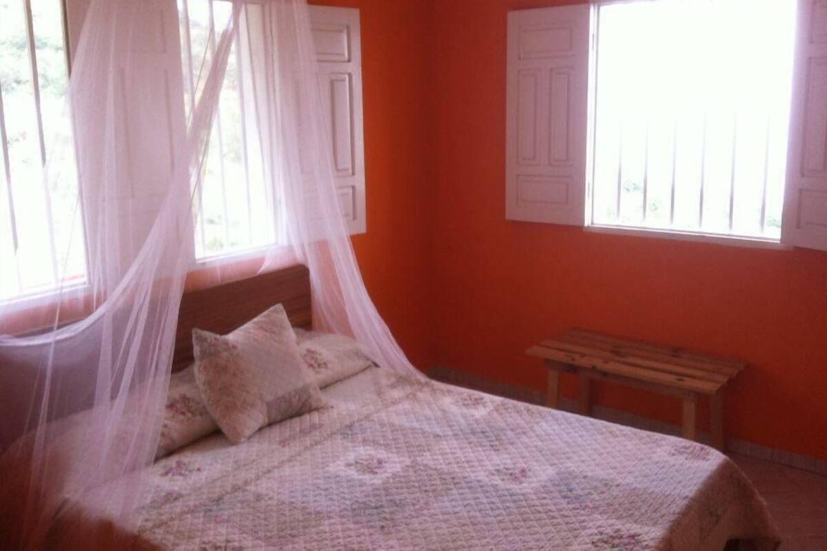 A bedroom with a bed, a bench, and a window with a white curtain.