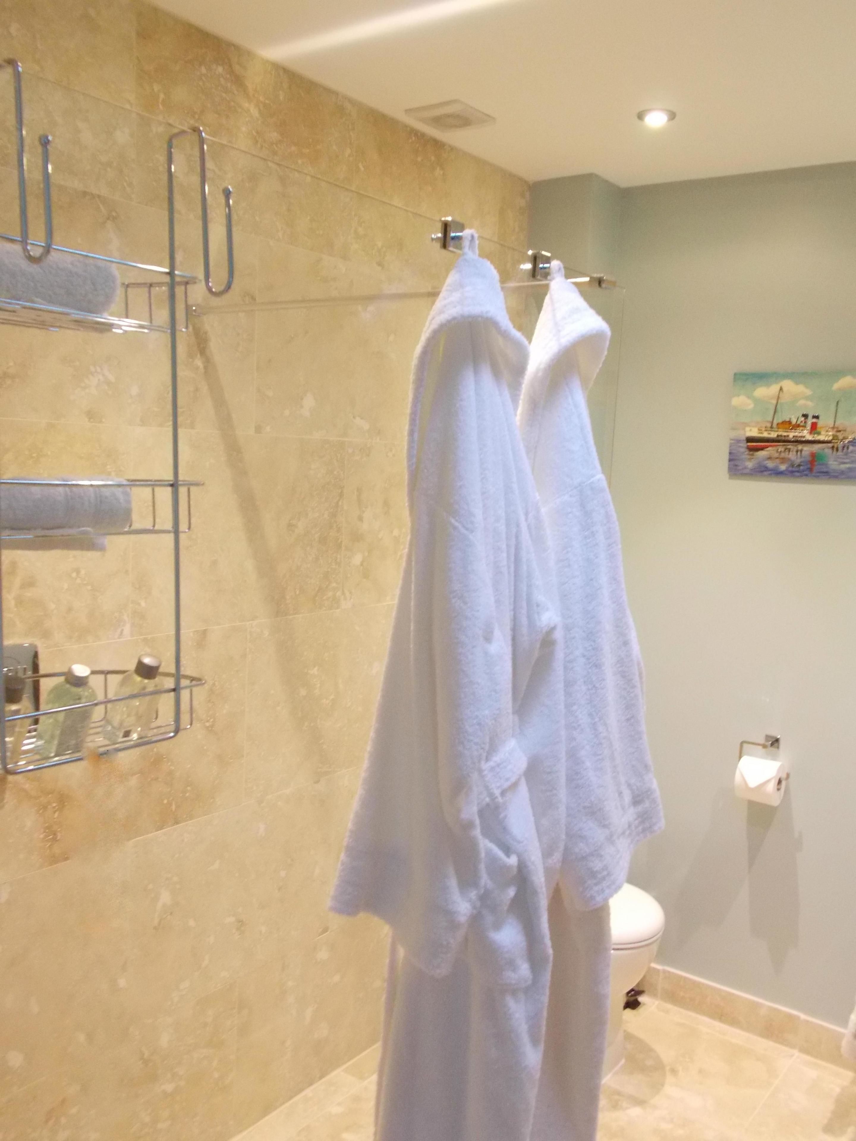 A bathroom with two white towels hanging on a rack, a glass shower caddy with toiletries, a painting on the wall, and a toilet with a paper towel holder.