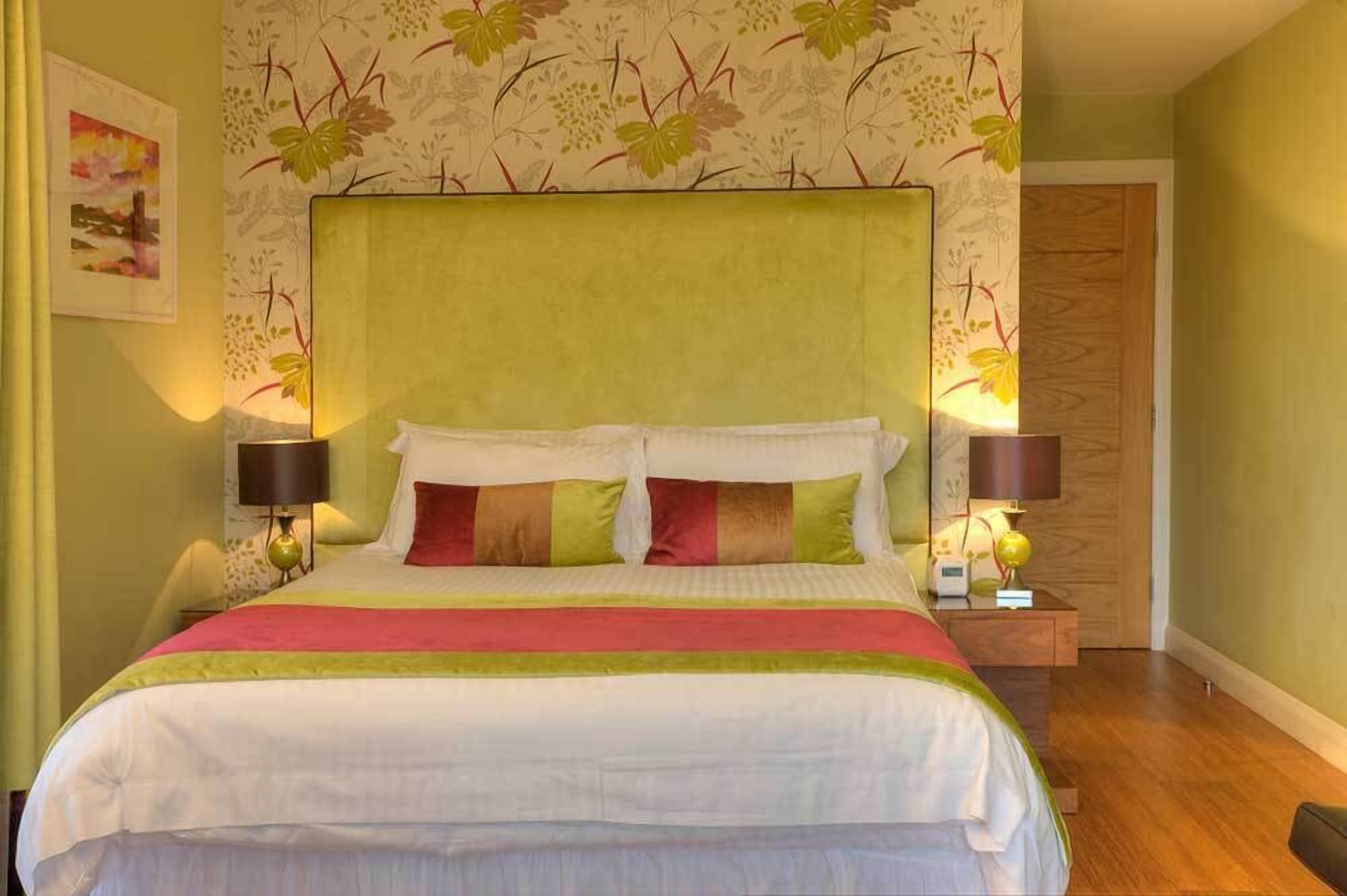 Een net opgemaakt bed met een groen hoofdbord, twee nachtlampjes en een behang met een bloemenmotief.