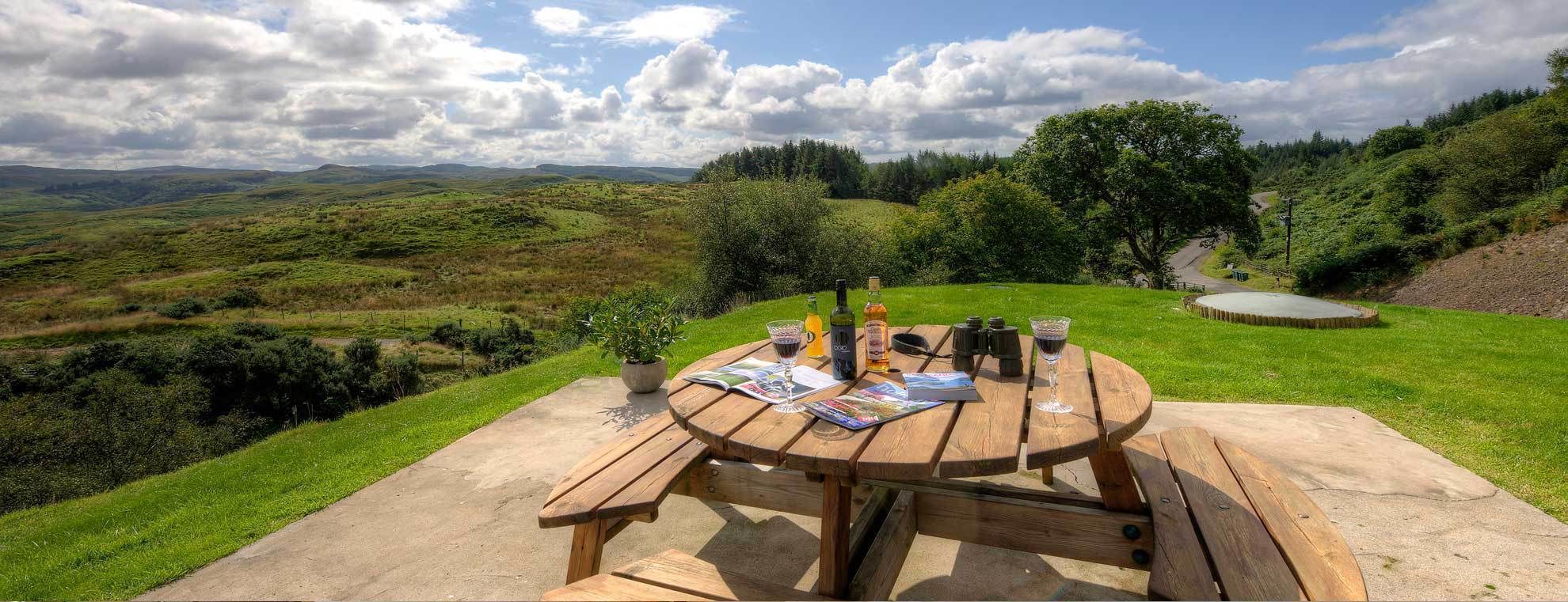 A picnic table with magazines, a bottle, and glasses on a grassy hill overlooking a valley.