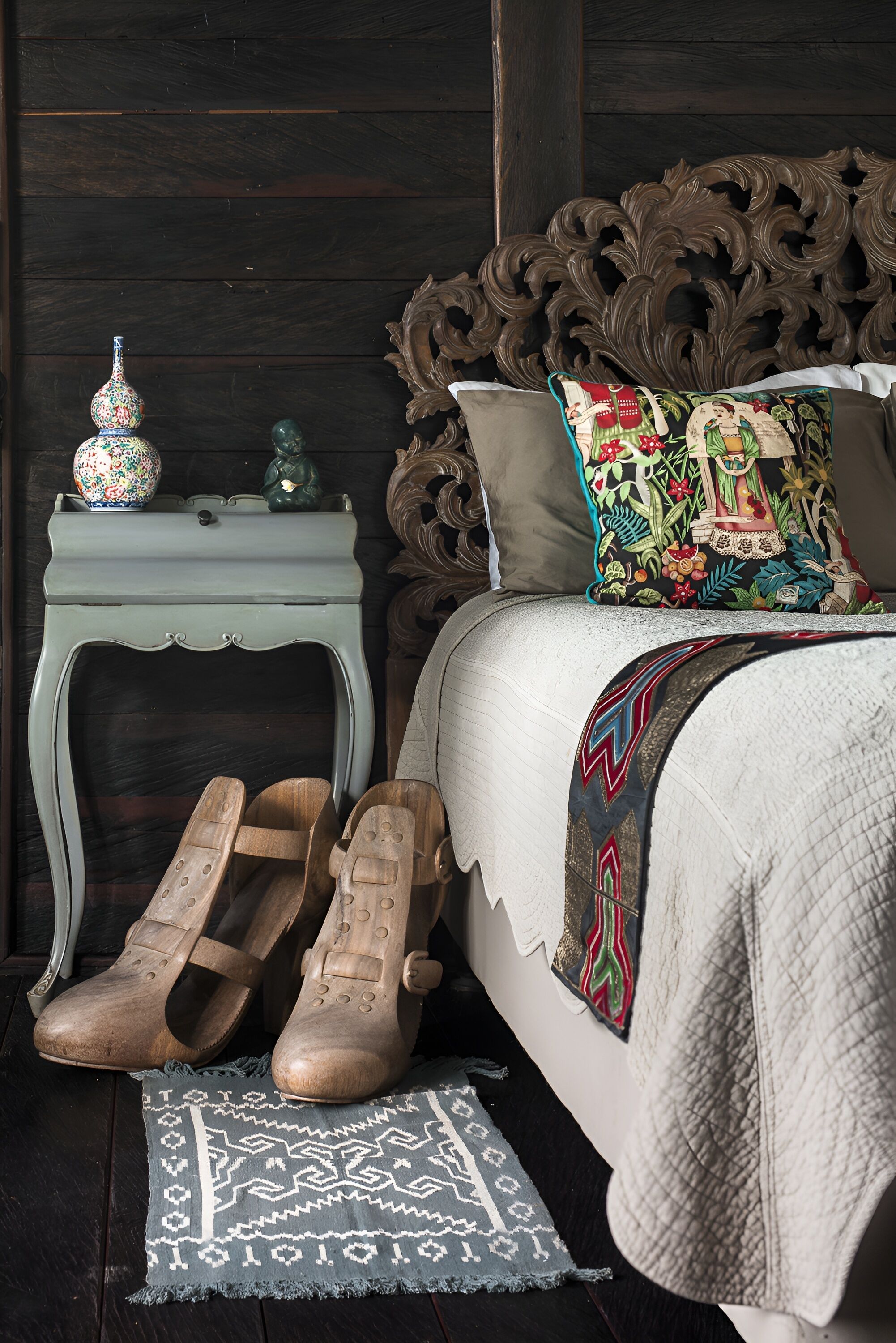 Ein Schlafzimmer mit hölzernem Kopfteil, einem Bett mit gemusterten Betttextilien, einem Nachttisch mit Dekoration und einem Paar Holzschuhe auf einem Teppich.