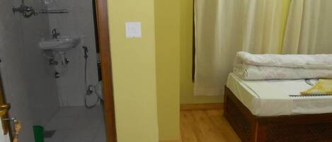 Una habitación con piso de madera, una cama con colchón y almohadas blancas, una toalla verde y un baño con lavamanos, espejo y ducha.