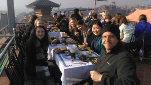 A group of people dining outdoors with a table set for a meal, a pagoda-like structure in the background, and a cityscape view. - Sunny Guest House and Cafe (Bhaktapur)