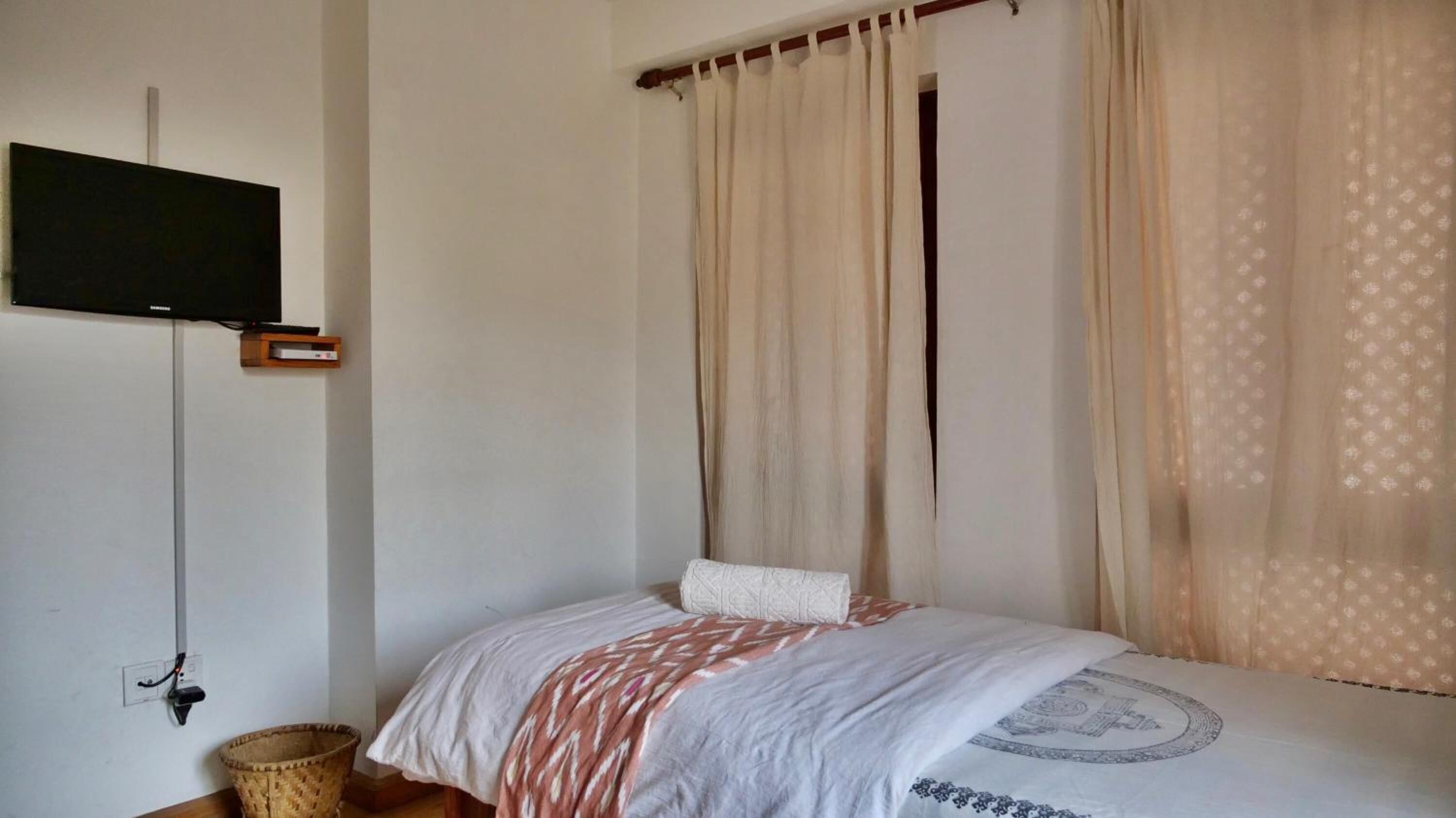 A bedroom with a bed, curtains, a television mounted on the wall, and a basket on the floor.