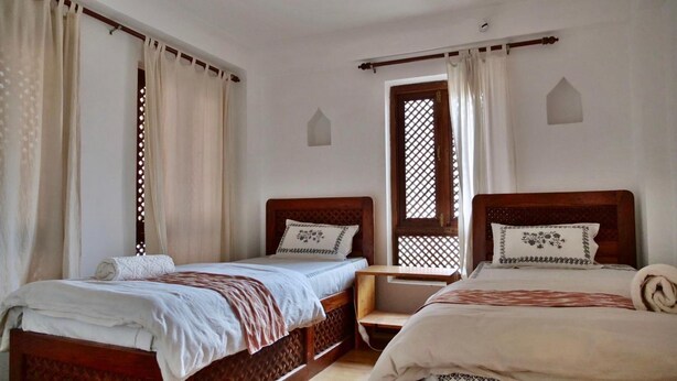 A room with two wooden beds, each with a headboard, white bedding, and a small bedside table. There is a window with a wooden lattice design and white curtains. - Sunny Guest House and Cafe (Bhaktapur)