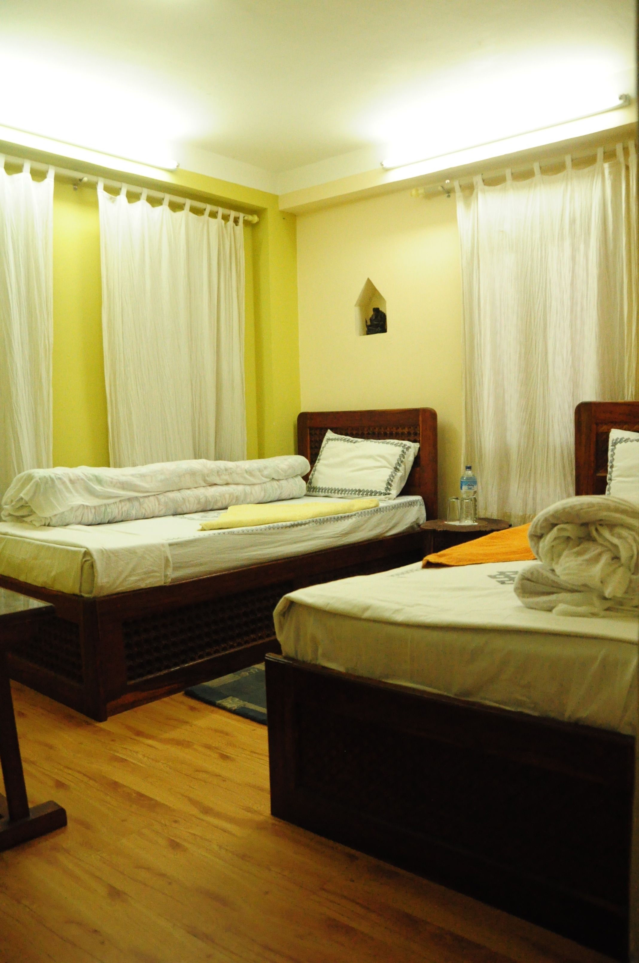 A hotel room with two single beds, a wooden floor, and a small wall-mounted nightstand with a water bottle.