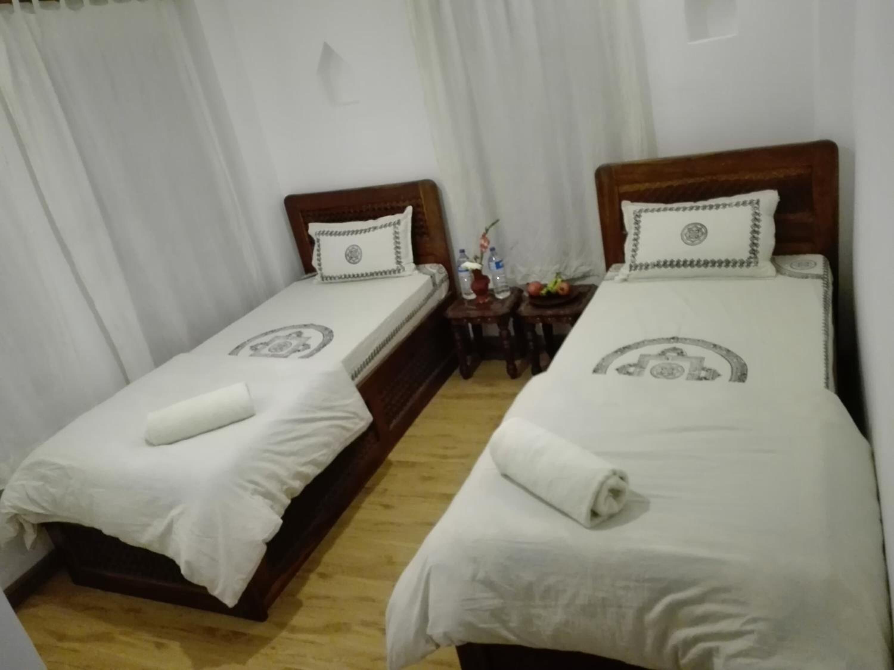 A hotel room with two single beds, each with a pillow and a rolled towel. There is a small table with a bottle and a bowl of fruit between the beds.