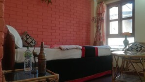 A room with a brick wall, a bed with a red and black bedspread, a wicker chair, a small table with a bottle and a vase, and a window with curtains. - Mountain Guest House (Bhaktapur)