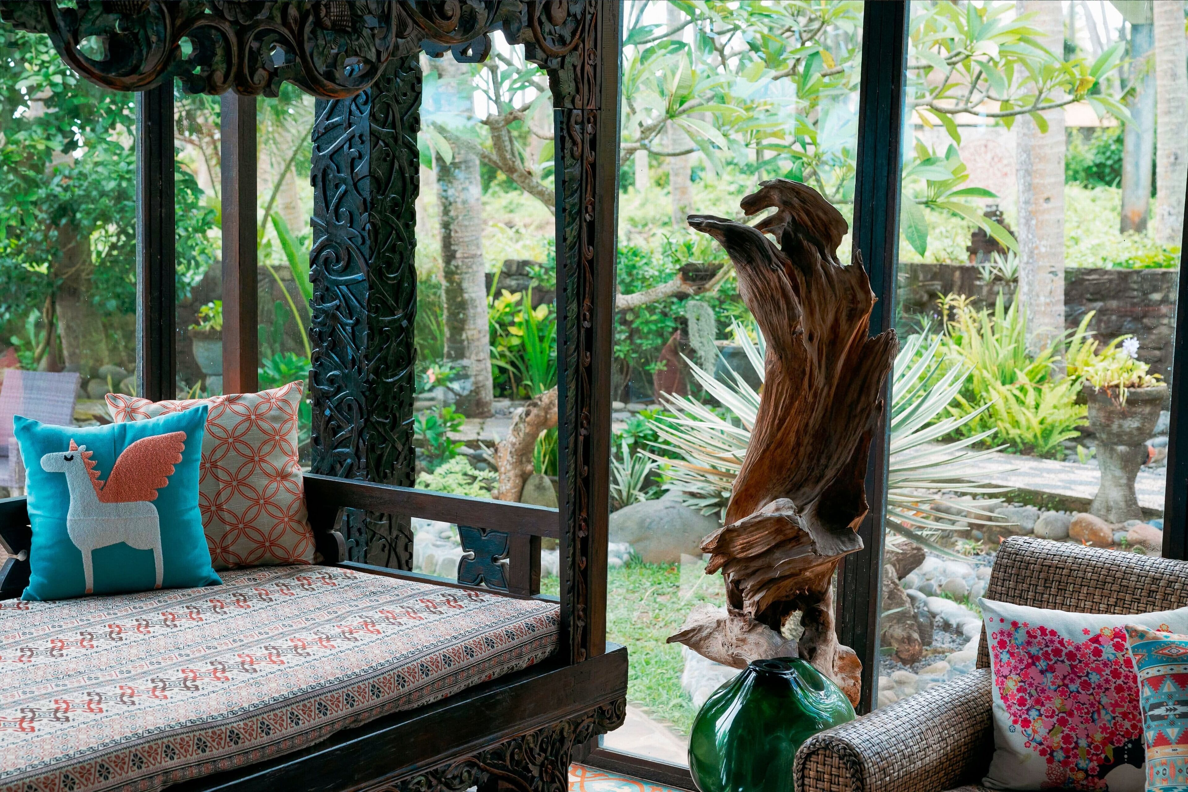 Une pièce avec un banc en bois, des coussins décoratifs et une vue sur un jardin, visible à travers une fenêtre ornée de motifs en métal.