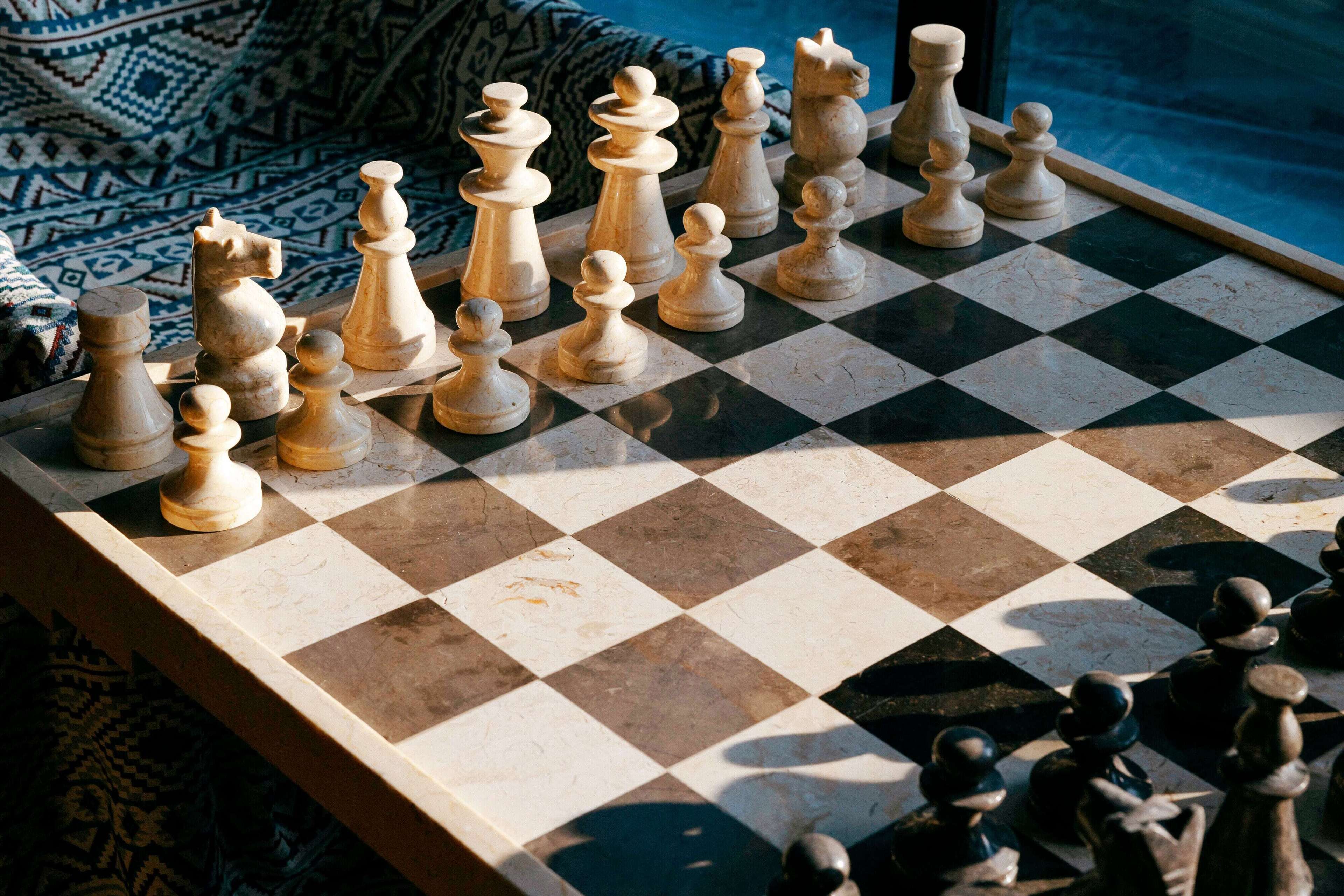 A chessboard with pieces set up for a game, placed on a table near a window with a patterned curtain.