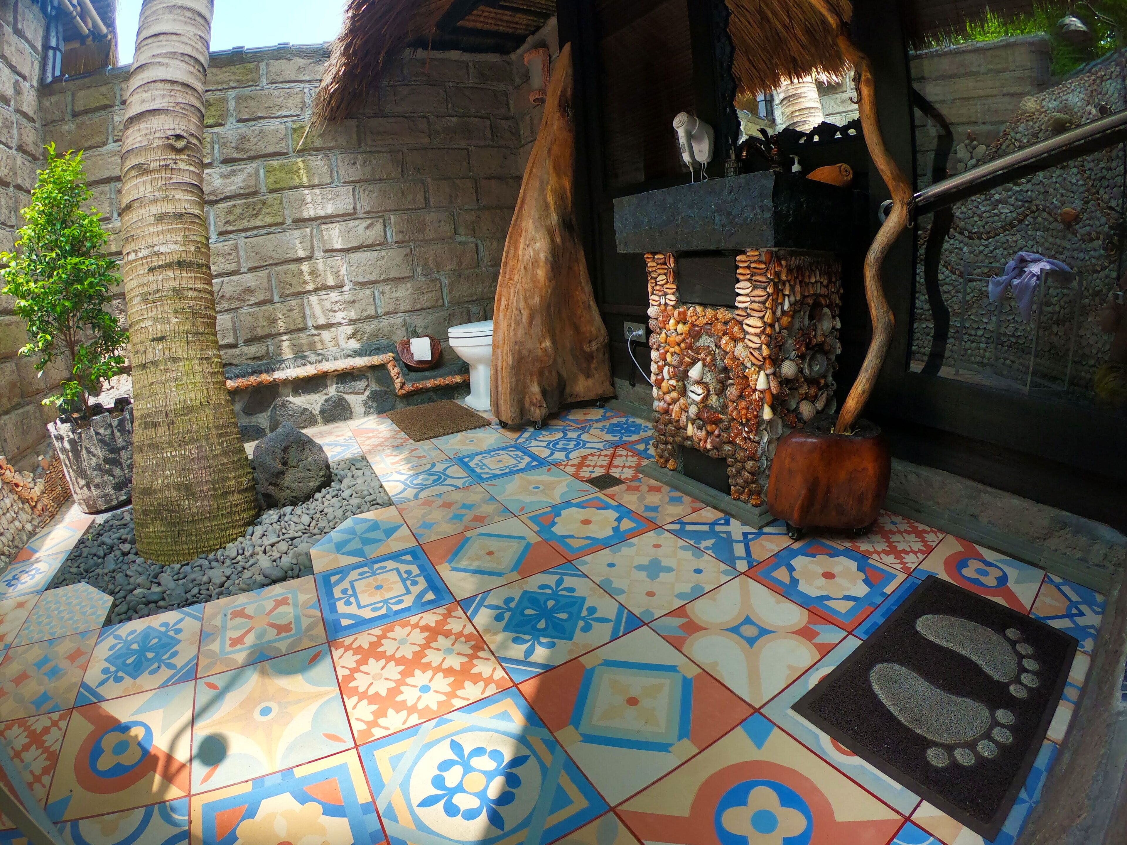 Een badkamer met een stenen muur, een betegelde vloer met geometrische patronen, een houten boomstam, een rustiek toilet en een plank met schelpen.