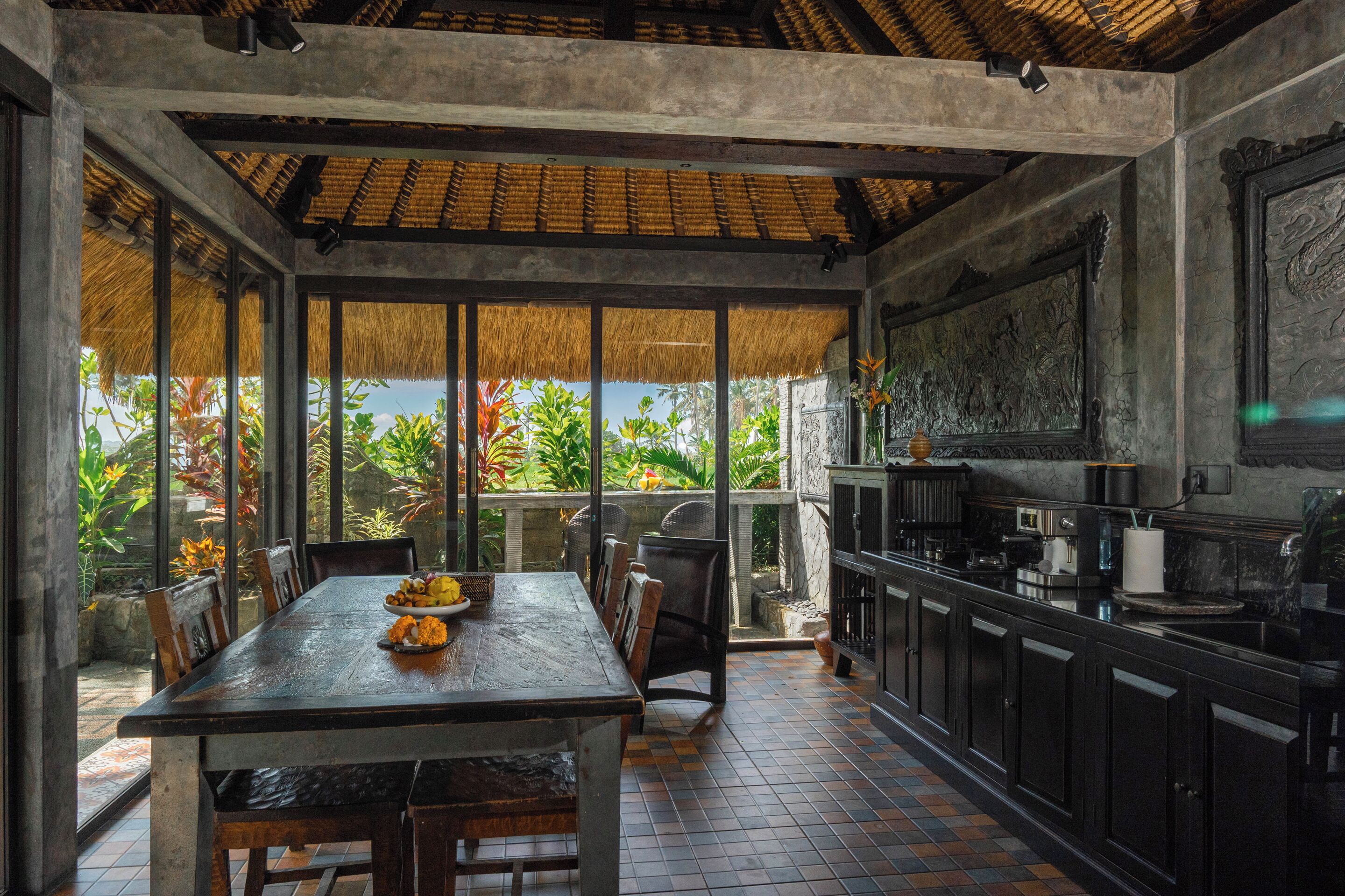 Un comedor con una mesa dispuesta para dos, una cocina con armarios oscuros y vistas a exuberante follaje a través de grandes ventanales.