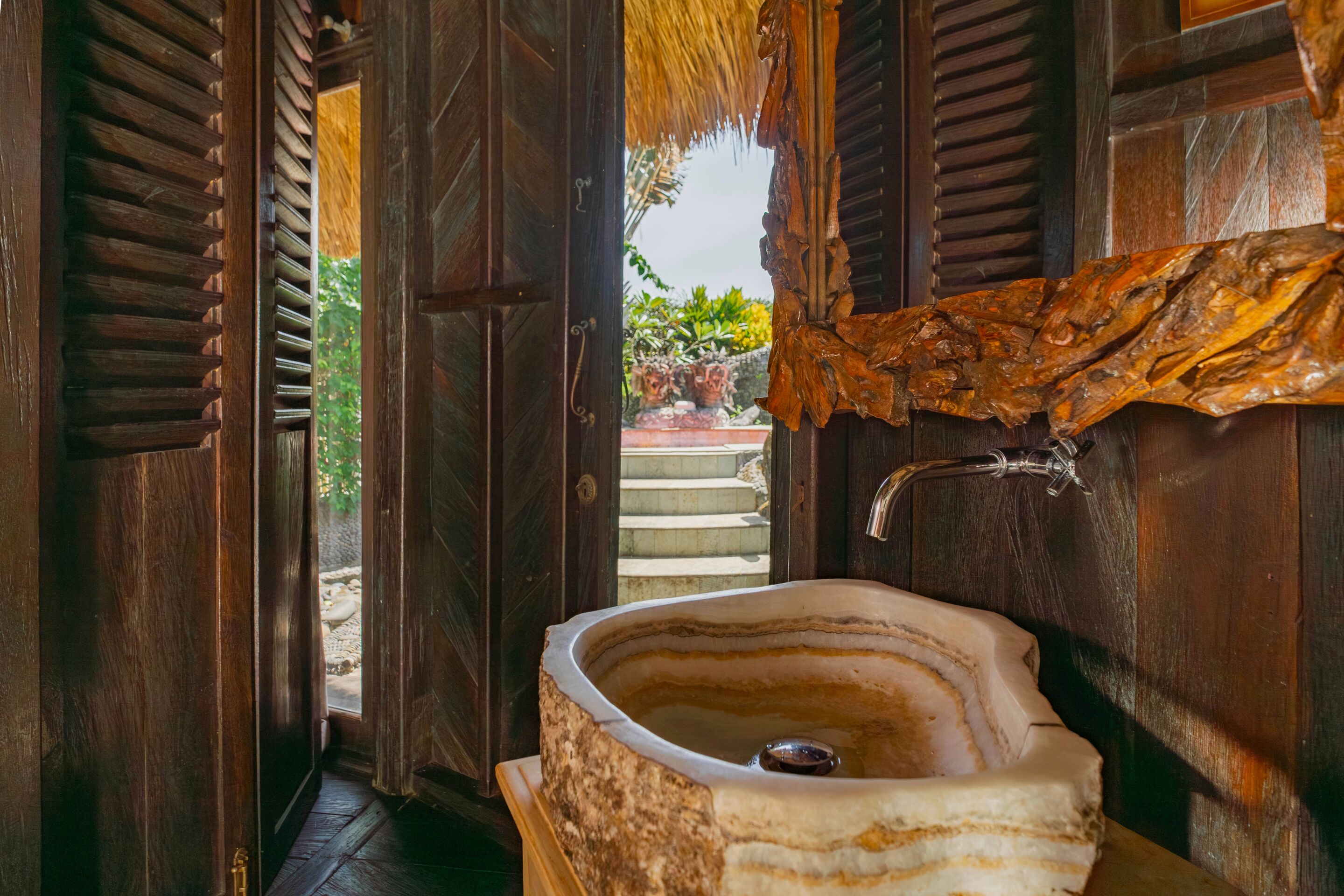 A bathroom with a large, natural stone sink, wooden doors, and a view of a garden outside.