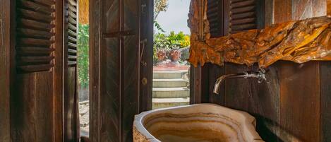 A bathroom with a large, natural stone sink, wooden doors, and a view of a garden outside.