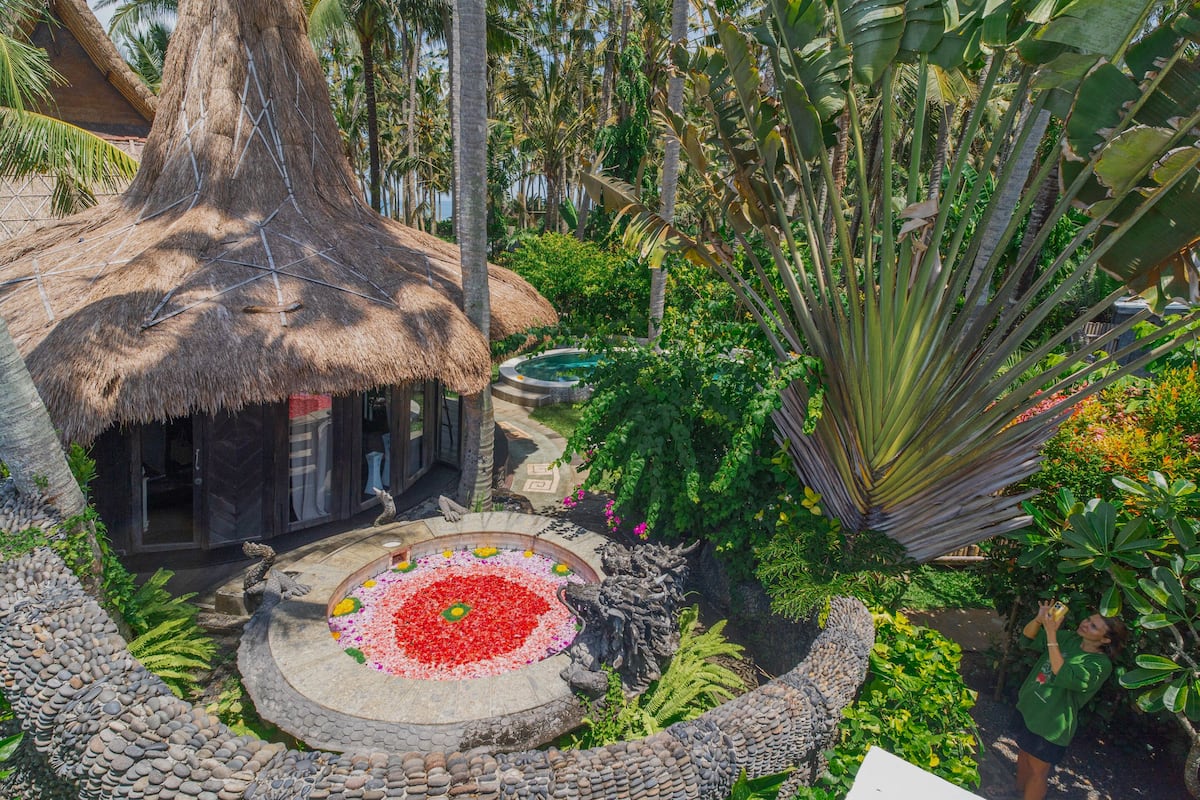 A thatched-roof structure surrounded by lush greenery and a circular water feature with red flowers.