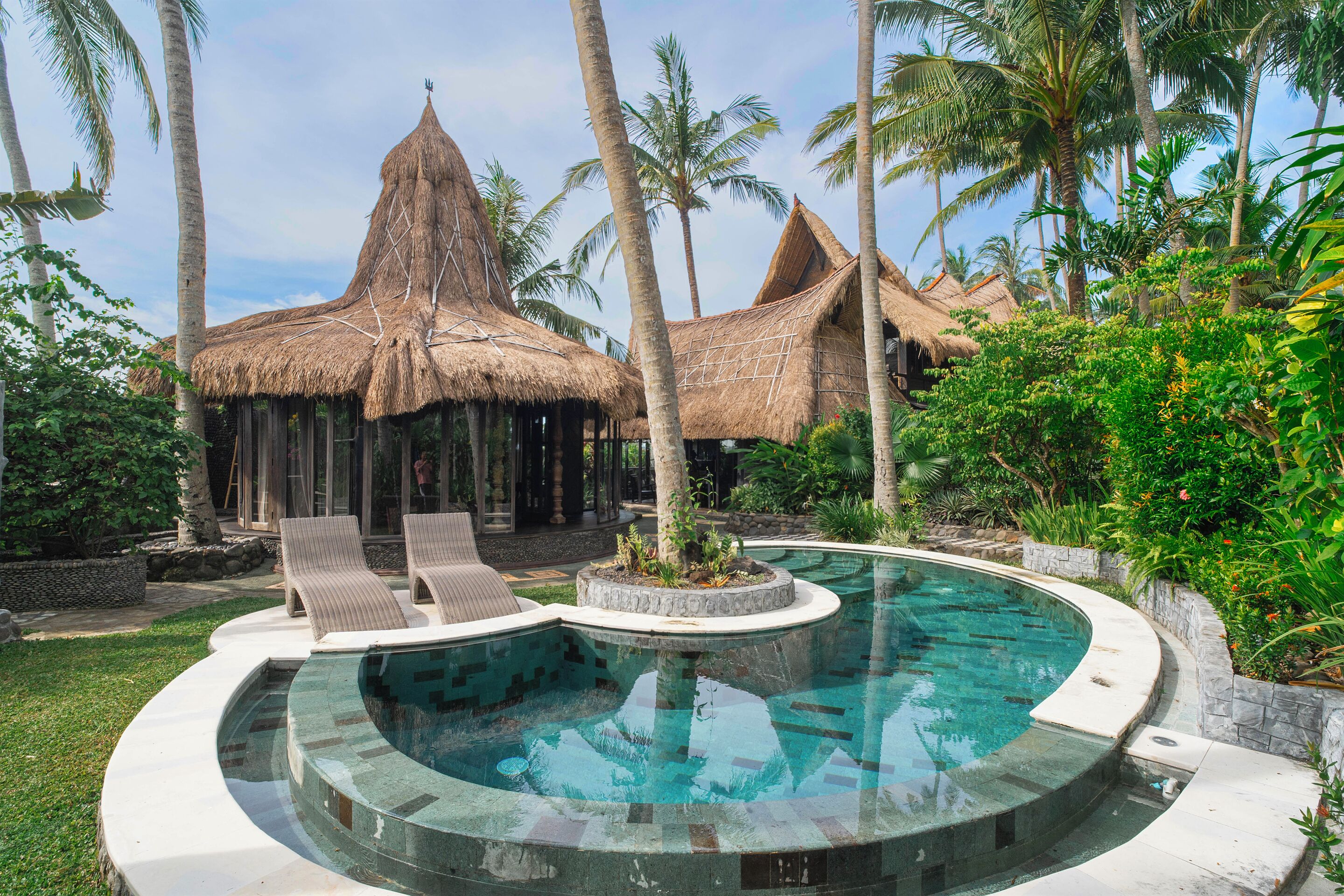 Ein Pool mit einer strohgedeckten Struktur im Hintergrund, umgeben von Palmen und Grünpflanzen.