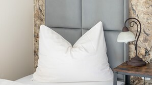 En seng med en hvid pude, et sengebord med en lampe og en væg med struktur.