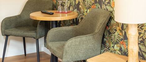 Una habitación con dos sillones tapizados en verde, una mesa de madera, una lámpara y un papel tapiz con motivos tropicales.