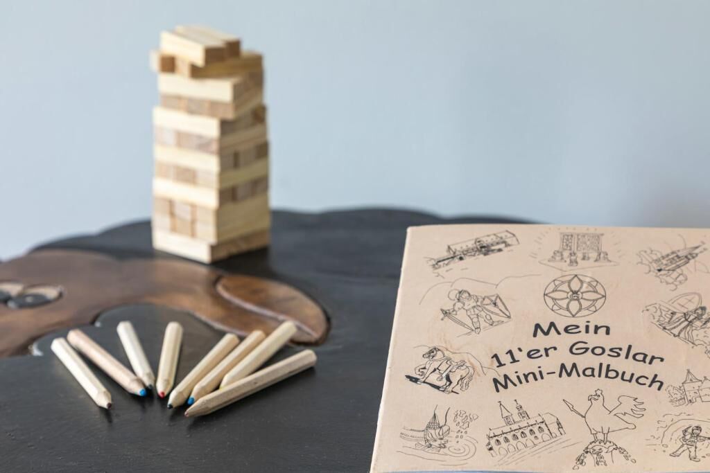 A book titled 'Mein 11'er Goslar Mini-Malbuch' with illustrations of various scenes, placed on a dark surface with wooden blocks and pencils nearby.