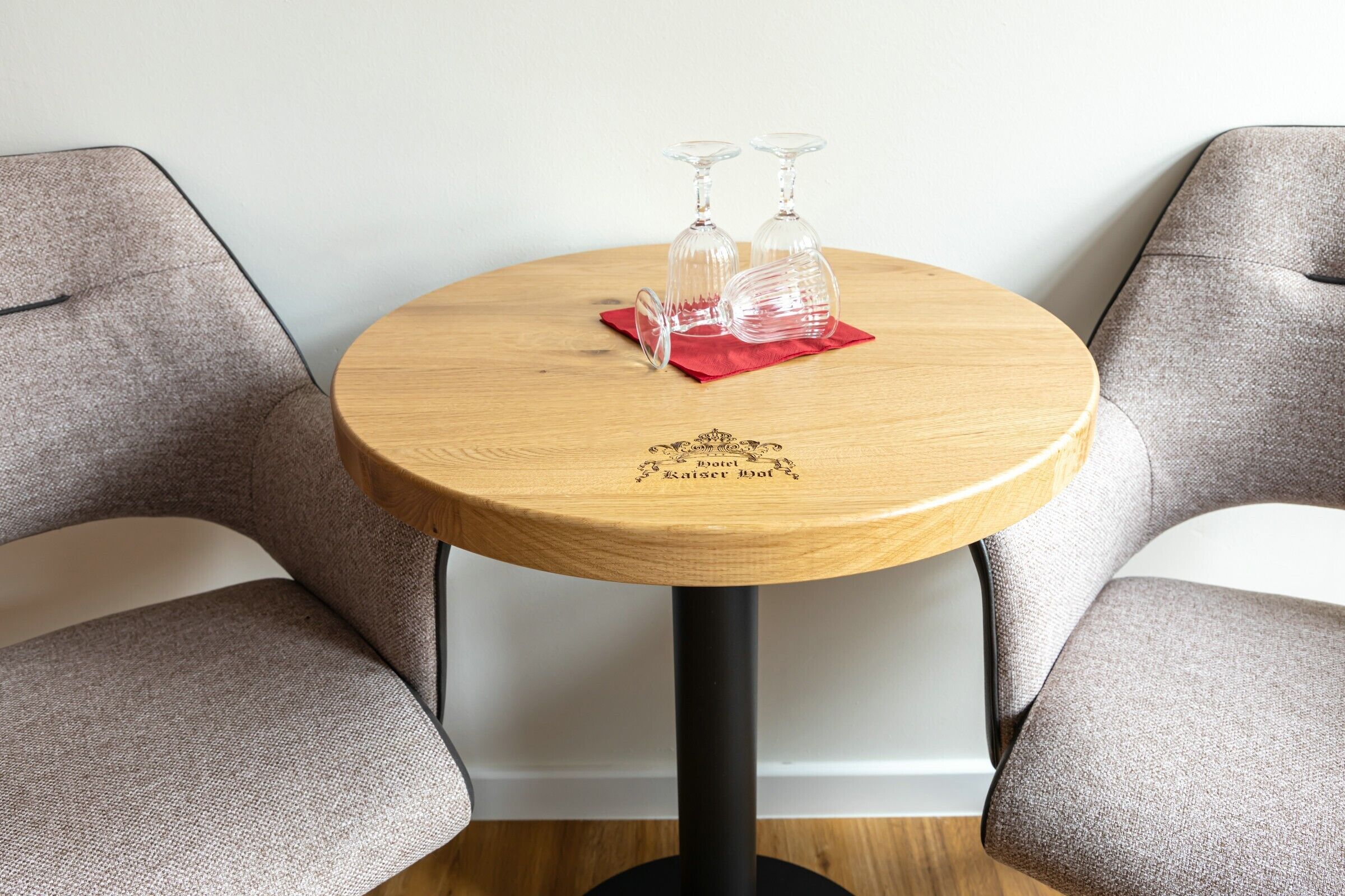 A round wooden table with two clear wine glasses on a red napkin, a logo, and two grey upholstered chairs.