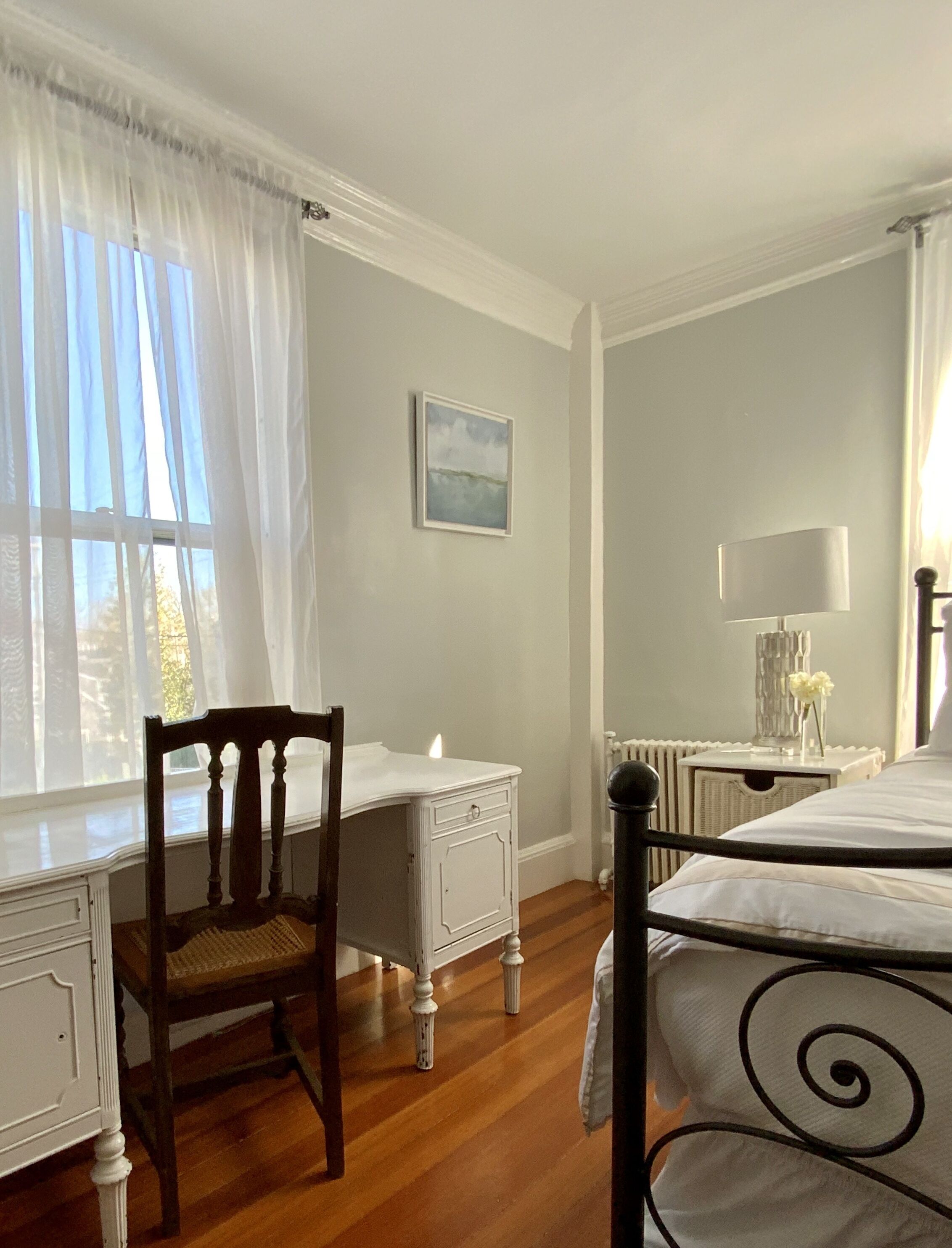Un dormitorio con una cama de madera, una silla, una mesita de noche con lámpara y una ventana con cortinas de gasa.