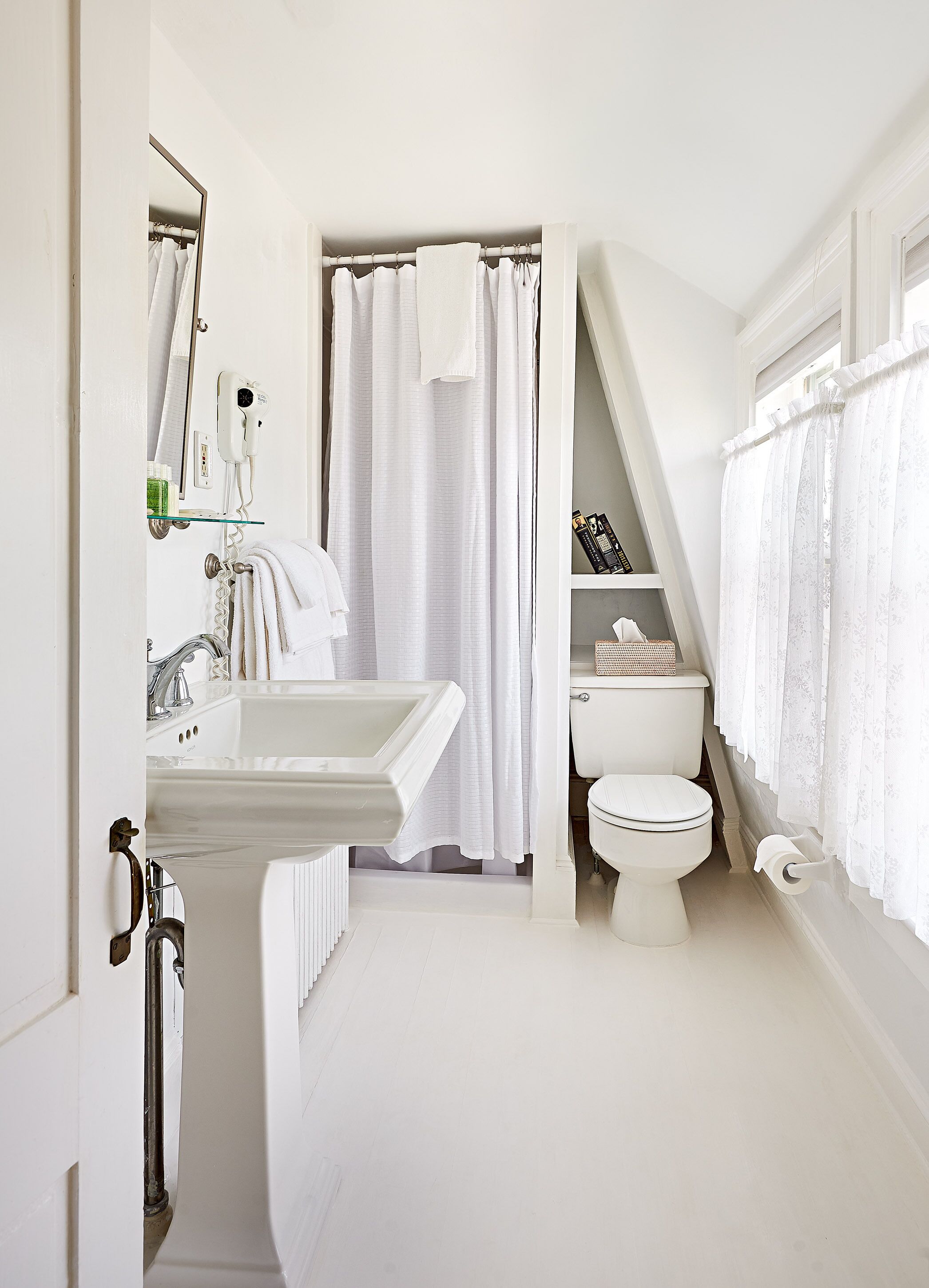 A bathroom with a white sink, toilet, and shower curtain. There is a glass shelf above the sink with toiletries.