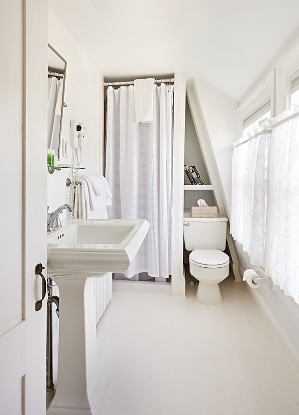 Un baño con lavamanos, inodoro y cortina de ducha blancos. Hay una repisa de vidrio sobre el lavamanos con artículos de tocador.